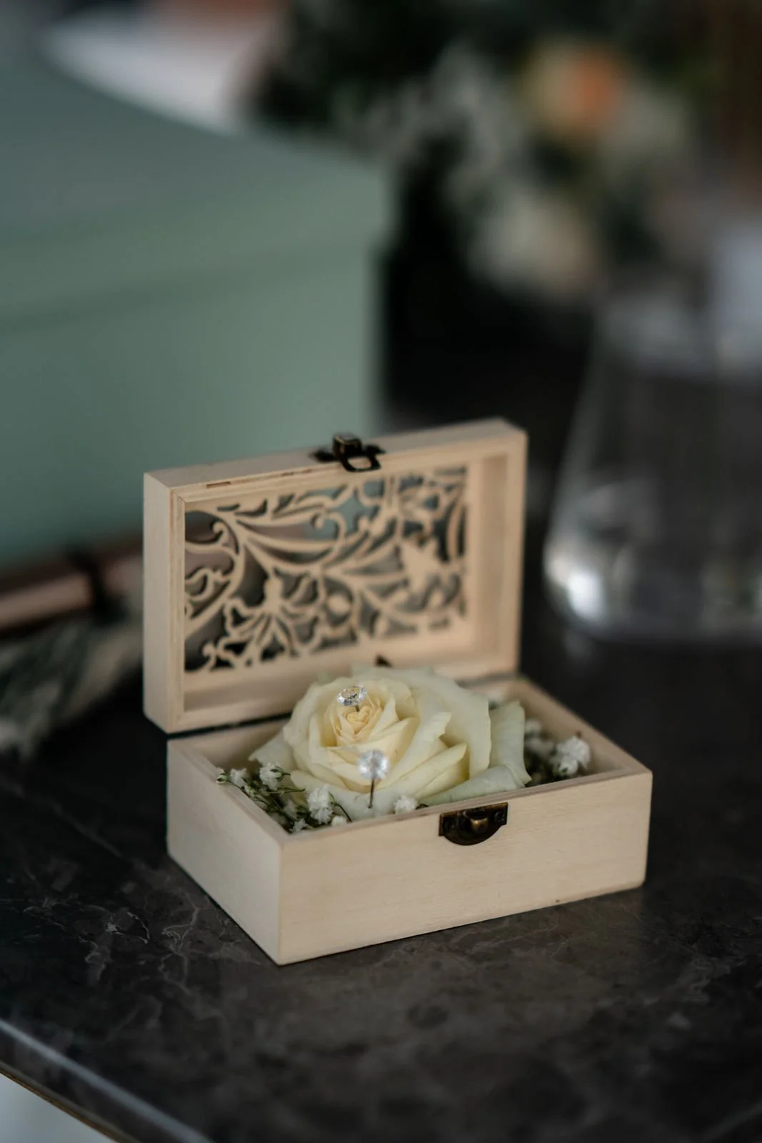 Eine offene Holzschatulle mit verschlüsselter Schnitzerei im Deckel, darin eine weiße Rose mit kleinen weißen Blumen und Diamanten