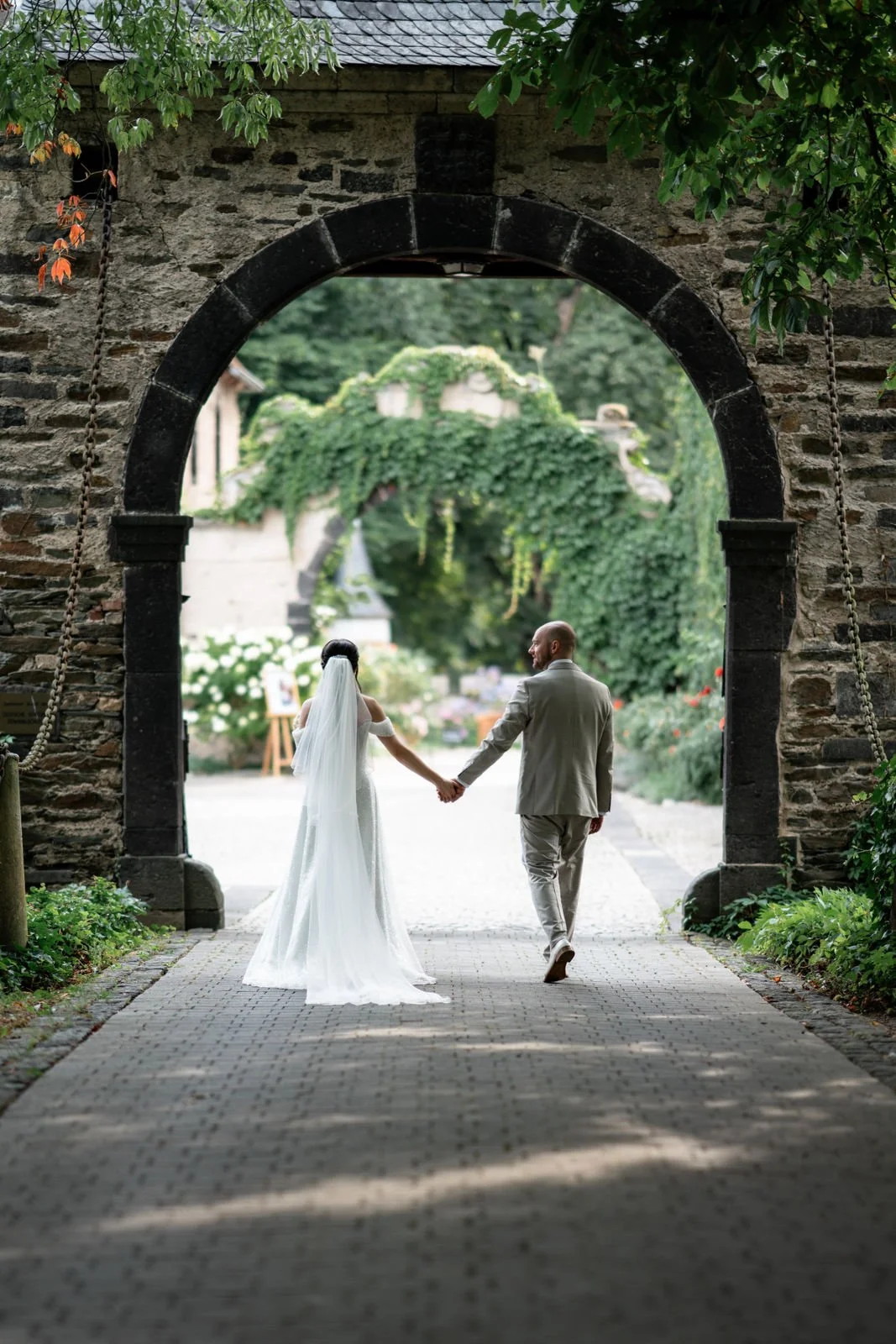 Ein Brautpaar hält Händchen und geht durch einen historischen Torbogen in einem Garten, umgeben von grünen Pflanzen und Blumen.
