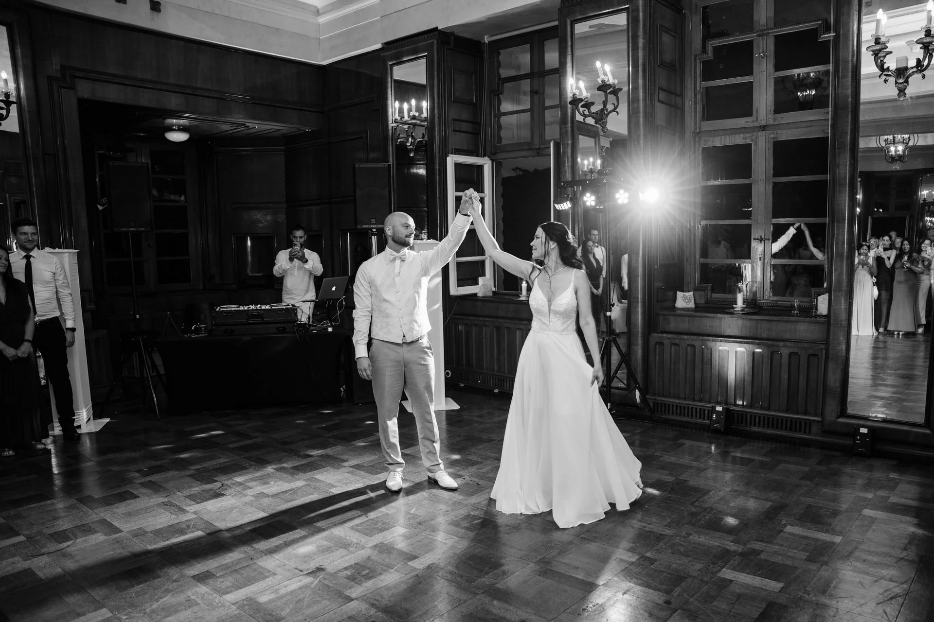 Braut und Bräutigam tanzen auf der Hochzeit, umgeben von Gästen, im eleganten Saal mit Holzvertäfelungen und großem Spiegel.