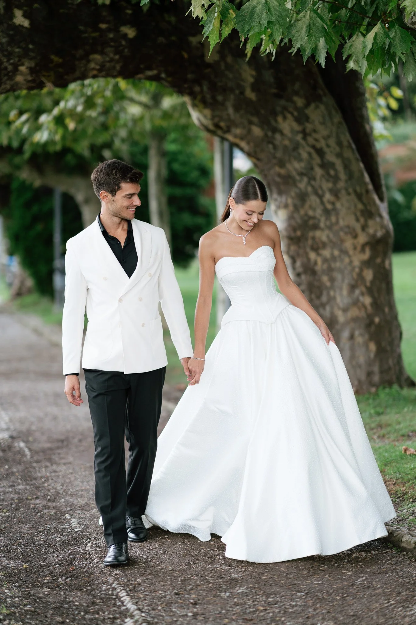 Ein Brautpaar hält Händchen am Comer See, die Braut trägt ein weißes Spitzenkleid und Schmuck, der Bräutigam einen weißen Anzug mit schwarzem Hemd, gehen unter einem großen Baum am Parkspaziert, lächelnd.