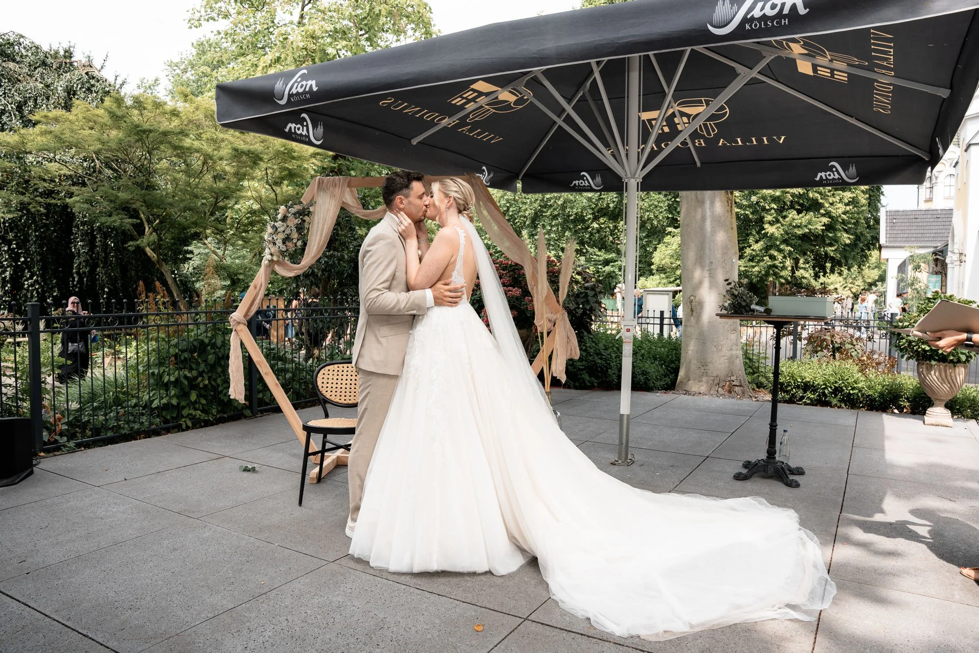 Ein Brautpaar küsst sich während ihrer Hochzeitszeremonie im Freien, unter einem großen schwarzen Sonnenschirm mit goldener Aufschrift, im Hintergrund grüne Bäume und ein Gartengewässer.