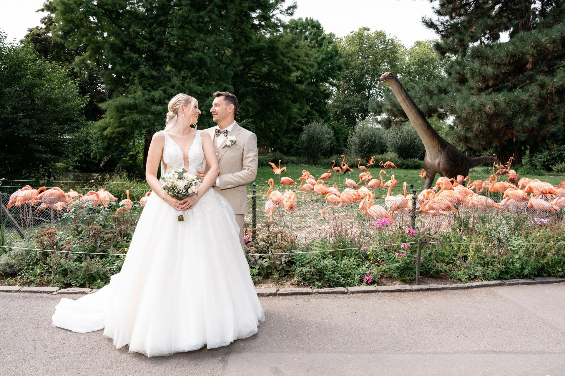 Ein Brautpaar in Hochzeitskleidung vor einer Flamingo- und Dinosaurierskulptur in einem Zoo oder Park, umgeben von Pflanzen und Bäumen.