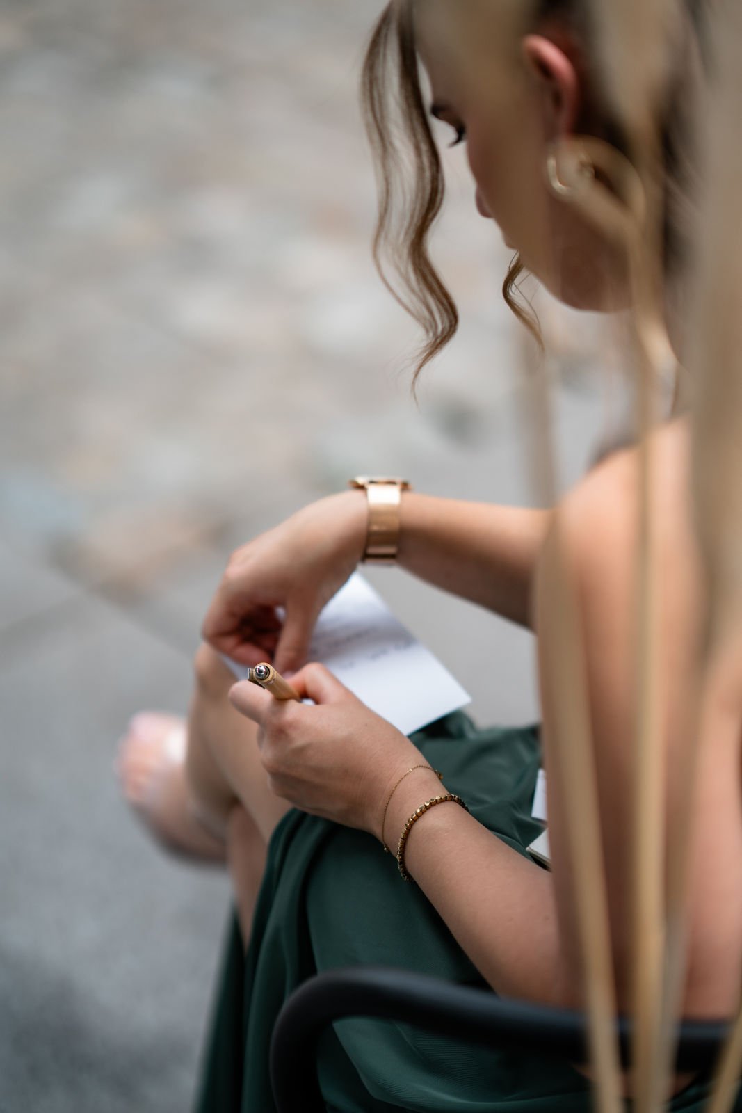 Eine Frau sitzt, schreibt auf einem Notizblock, trägt eine goldene Uhr, eine goldene Armbanduhr und eine kleine Goldkette, mit lockigem Haar und Ohrringen.