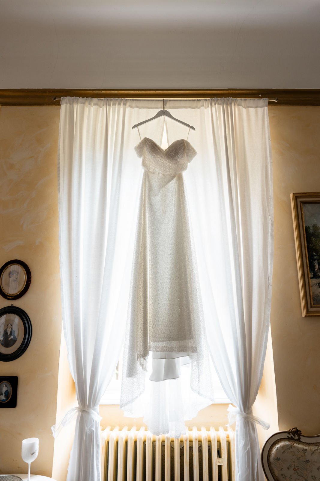 Ein Hochzeitskleid hängt an einem Kleiderbügel vor einem weißen Fenster mit Vorhängen, die durch das Licht hinter dem Kleid erleuchtet werden.