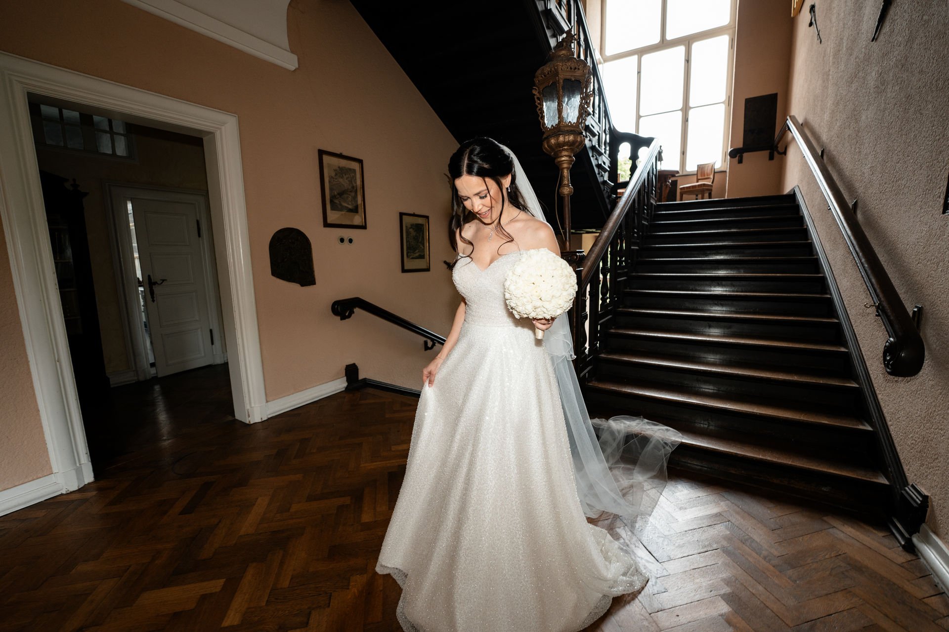 Braut in Brautkleid hält einen Brautstrauß und lächelt in einem Flur mit dunklem Holztreppe und großen Fenstern im Hintergrund.