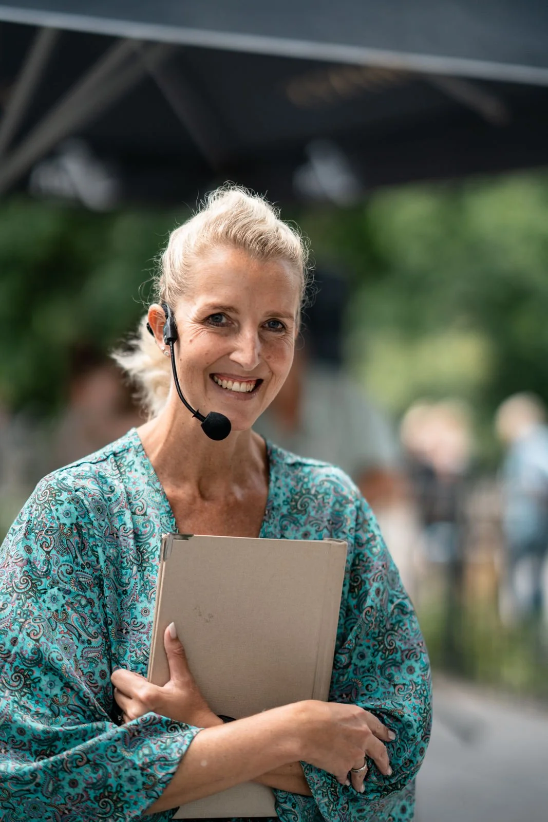 Eine lächelnde ältere Frau trägt ein Headset und hält ein beigeordner gefaltet in den Armen vor ihrem Körper, während sie draußen steht.