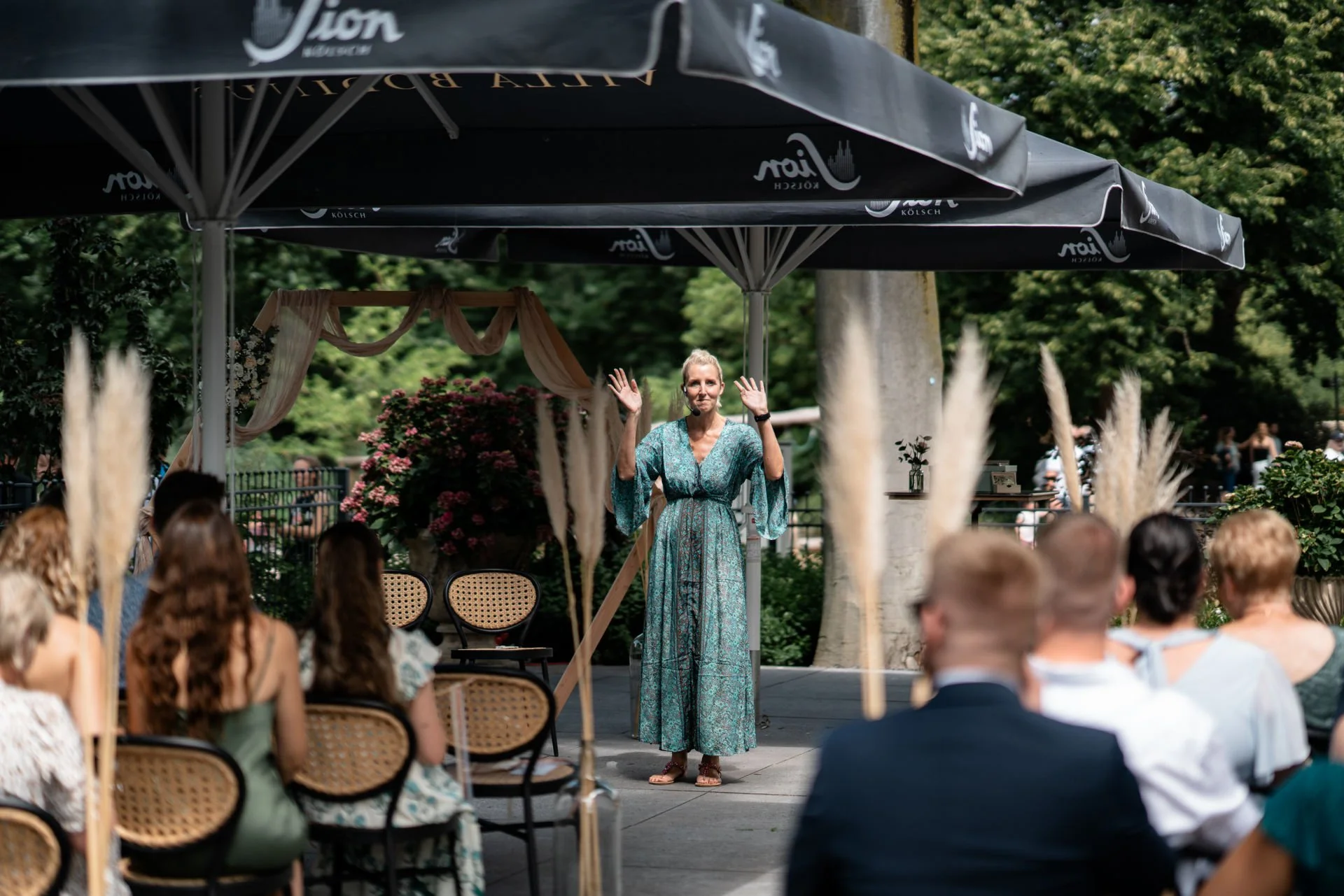 Eine Frau steht bei einer Zeremonie im Freien unter einem großen Sonnenschirm. Sie trägt ein langes, grünes Kleid mit Mustern und hat die Hände erhoben. Im Vordergrund sitzen mehrere Menschen auf Stühlen, die einer Ansprache oder Präsentation zuhören