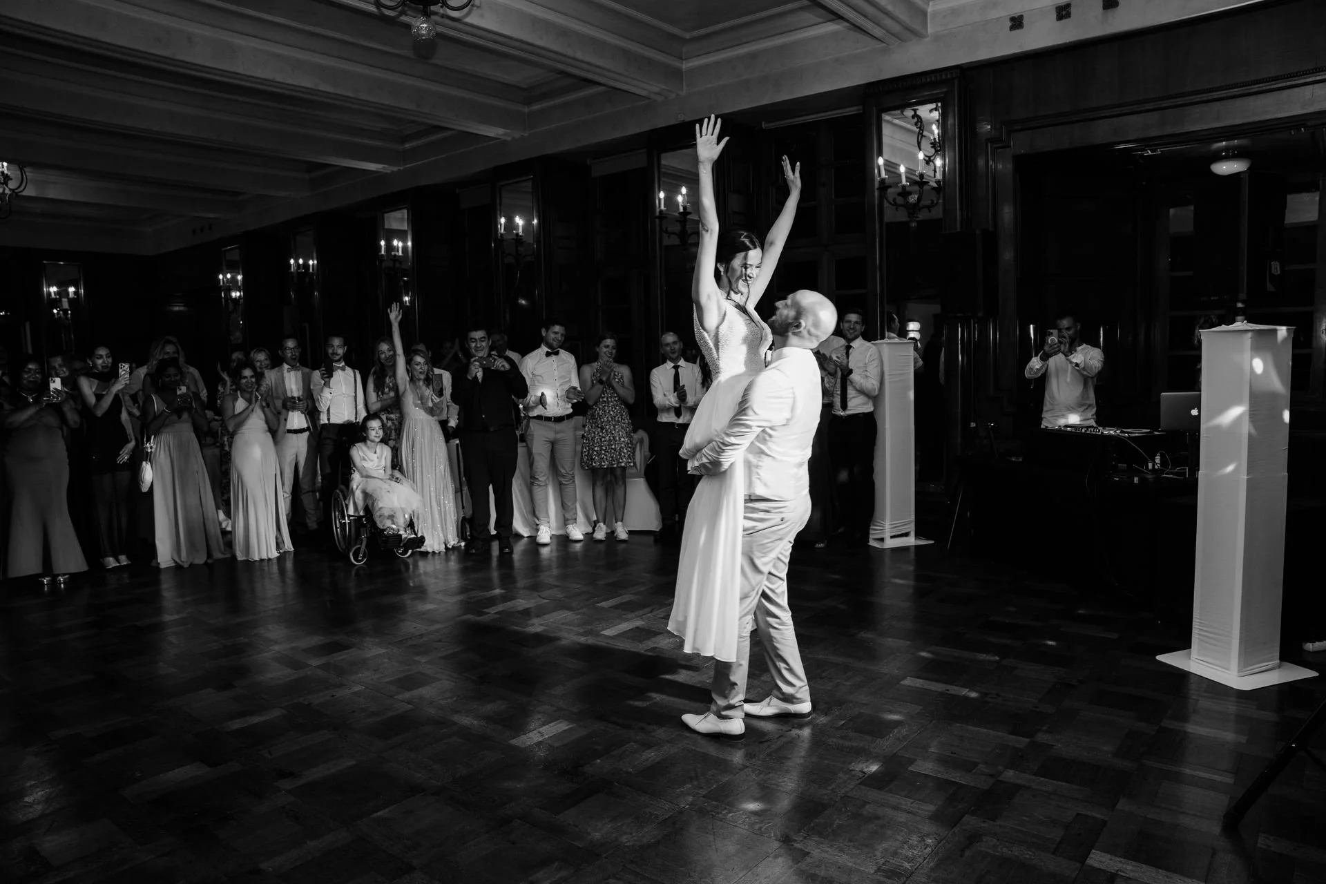 Ein Brautpaar tanzt bei einer Hochzeit, während die Gäste im Hintergrund zuschauen, einige mit Handys. Das Gesicht der Braut ist sichtbar, während der Bräutigam sie küsst, beide im weißen Outfit. Die Umgebung ist eine elegante, dunkle Holztapete mit 