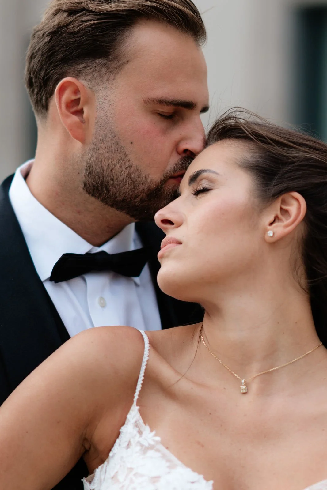 Ein verliebtes Paar in eleganter Kleidung, der Mann trägt einen schwarzen Anzug mit Krawatte, die Frau ein weißes Kleid mit Spitze und Schmuck, teilt einen intimen Moment im Freien.