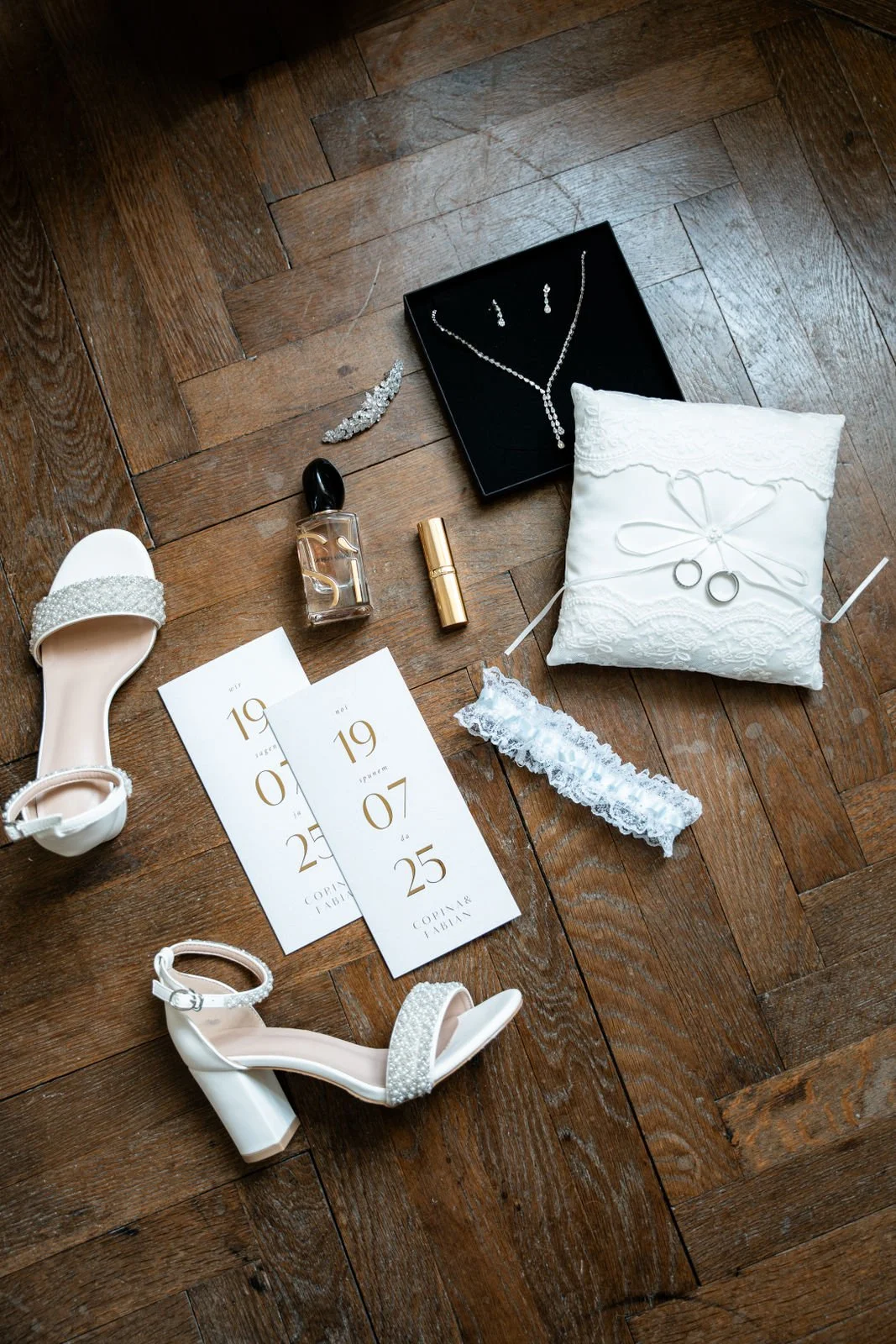 Hochzeitsaccessoires und Einladungskarten auf einem Holzboden, darunter weiße Schuhe, Schmuck, Parfüm, Lippenstift, Hochzeitskarten, Ringe, Lavendelband, Stickereiband und ein Brautkissen.