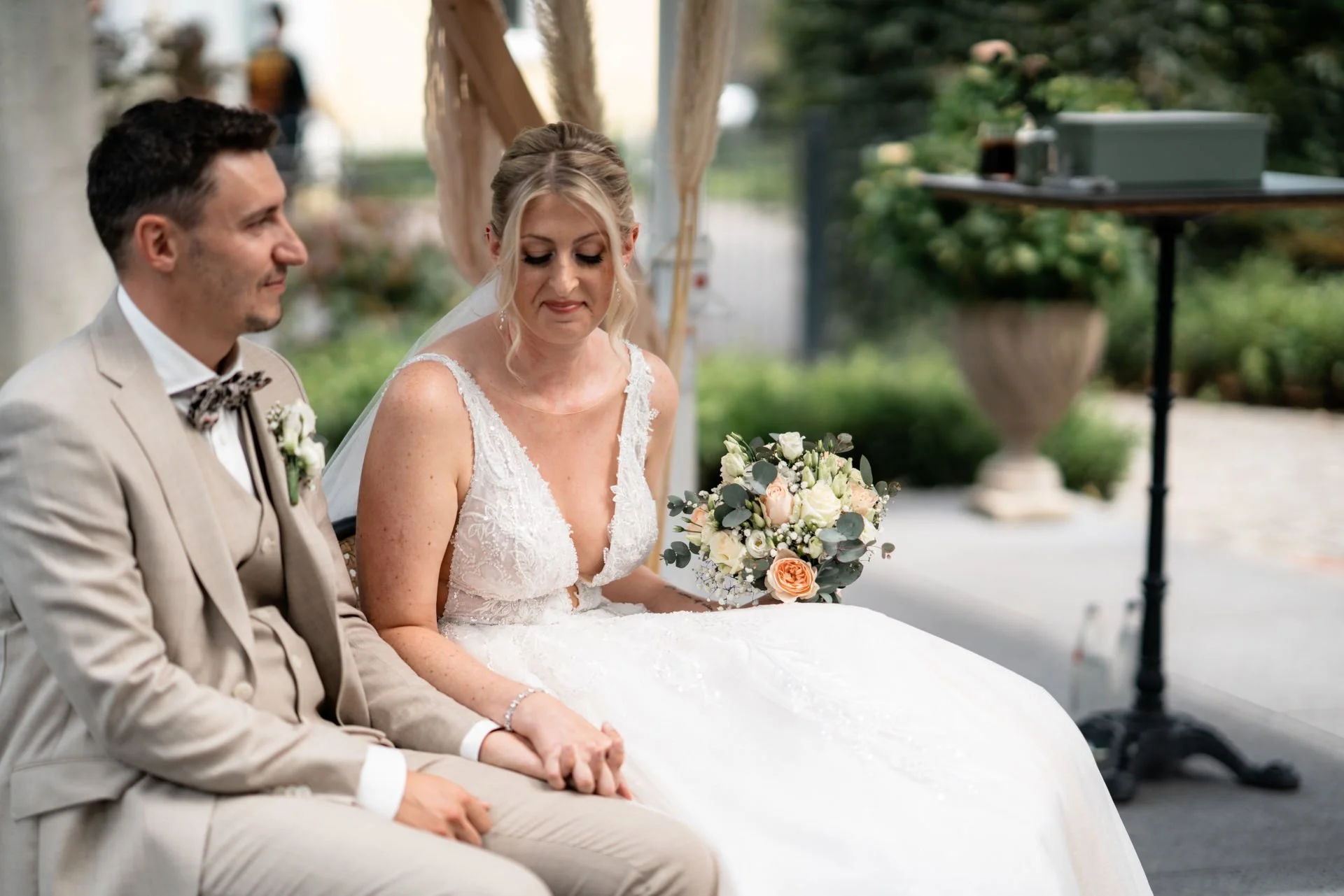 Braut und Bräutigam sitzen bei ihrer Trauung, die Braut hält einen Blumenstrauß, beide sind elegant gekleidet, im Hintergrund sind Pflanzen und Dekorationen sichtbar.