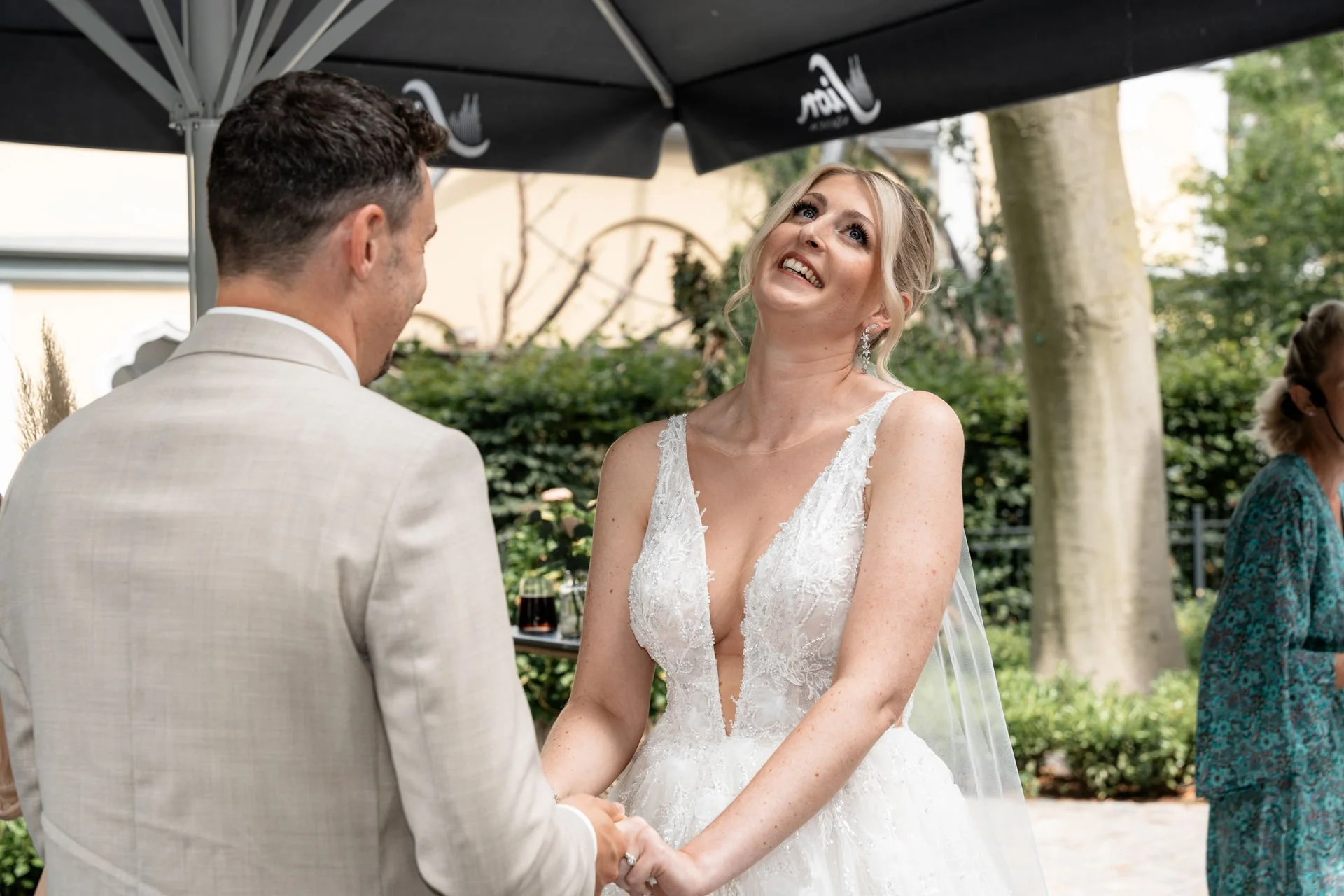 Braut und Bräutigam während ihrer Zeremonie im Freien, die Braut trägt ein weißes Hochzeitskleid mit V-Ausschnitt, der Bräutigam trägt einen hellen Anzug, im Hintergrund grüne Bäume.