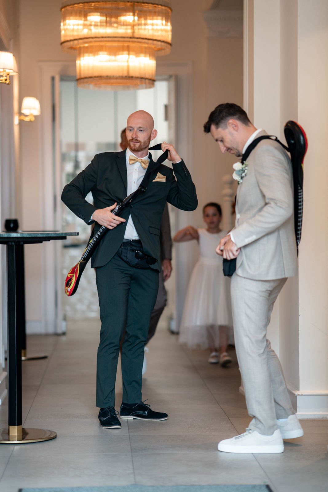 Zwei Männer in Anzügen mit Golfschlägern, der eine mit schwarzem Anzug und goldener Fliege, der andere in einem hellen Anzug, im Hintergrund ein junges Mädchen in weißem Kleid.