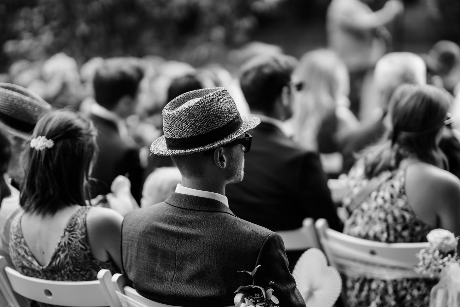 Menschen bei einer Hochzeit im Freien, sitzt in Reihen, einige tragen Hüte und Sonnenbrillen, in Schwarz-Weiß-Fotografie.