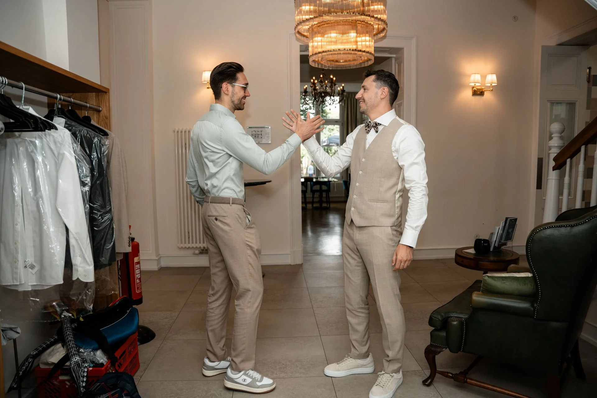Zwei Männer begrüßen sich mit Handshake in einem eleganten Raum, umgeben von Kleidung und Möbeln.