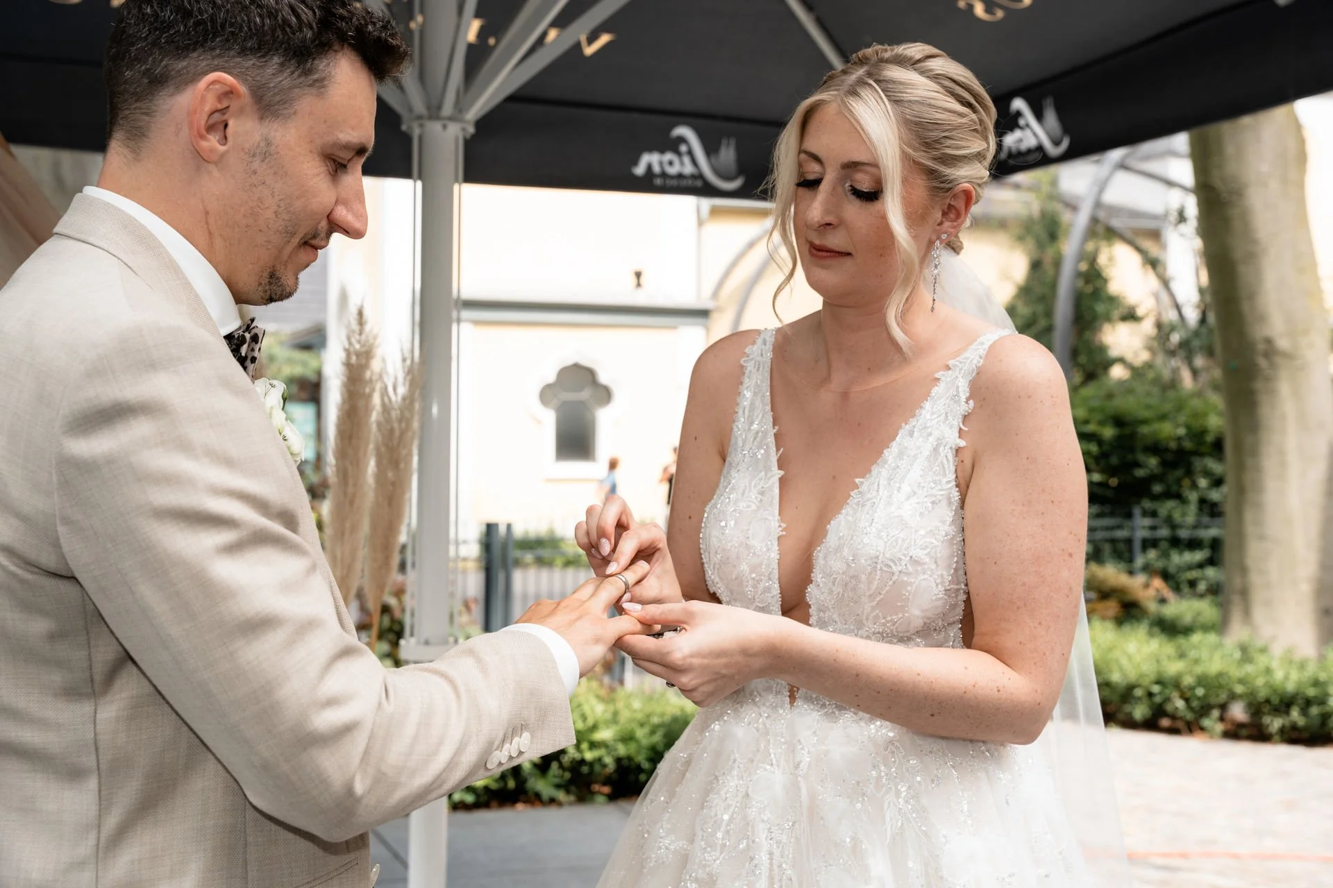 Ein Brautpaar bei der Eheschließung im Freien, der Bräutigam steckt der Braut den Ehering an, beide tragen formelle Hochzeitskleidung, im Hintergrund befindet sich eine offene Landschaft mit Bäumen.