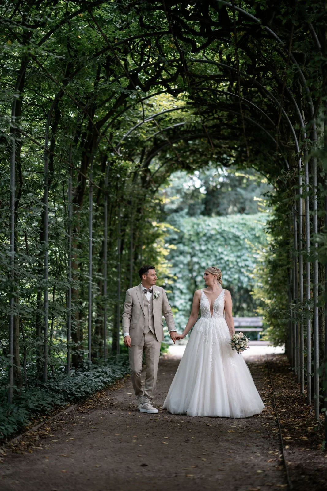 Ein Brautpaar hält sich an Händen in einem begrünten Park unter einem gewölbten Wäsche, trägt formelle Hochzeitskleidung, die Braut in einem weißen Kleid mit V-Ausschnitt und der Bräutigam in einem beige Anzug, während sie sich ansehen.