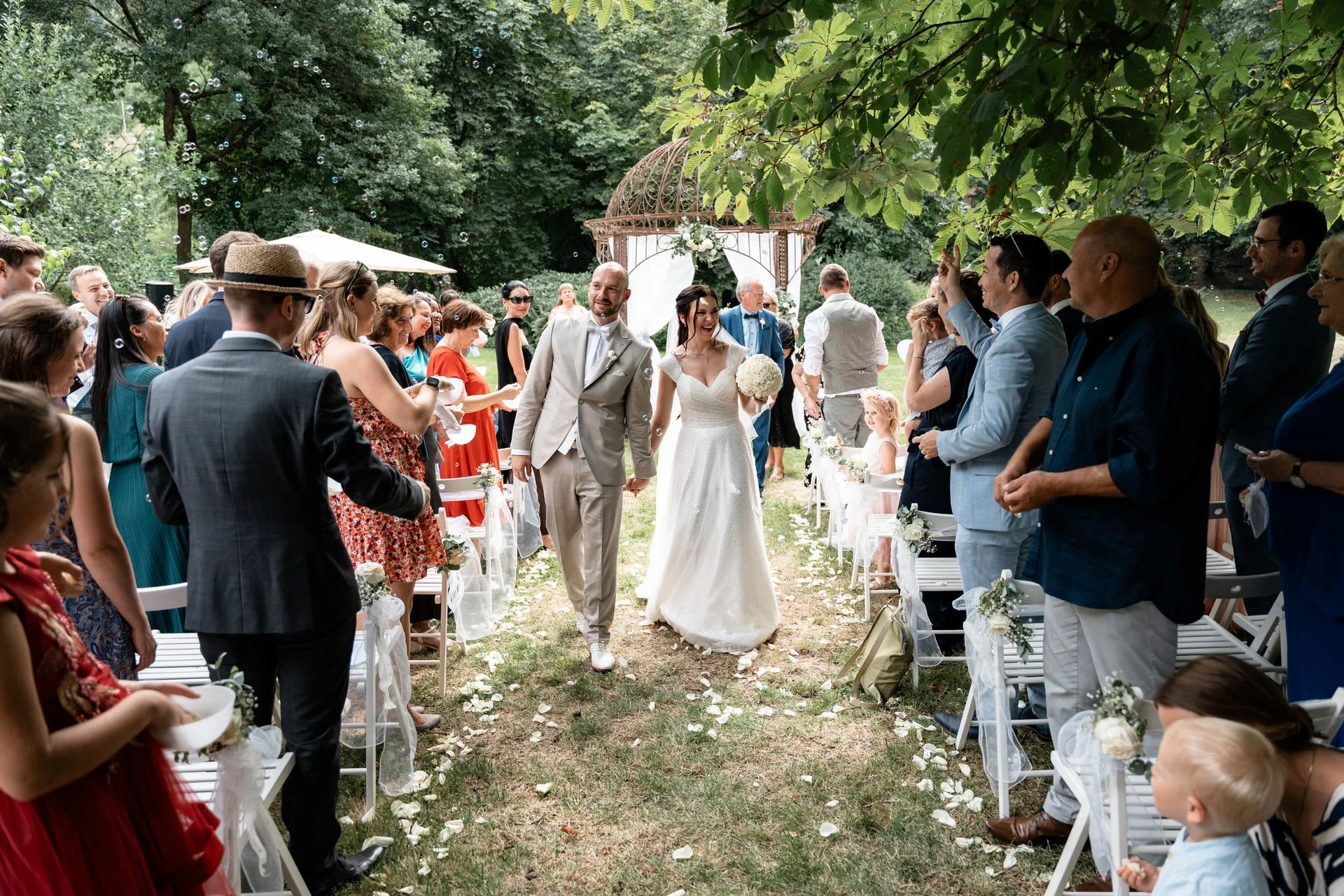 Ein Hochzeitspaar läuft Hand in Hand auf einer Gartenwiese, umgeben von Gästen, die sie begrüßen und feiern. Im Hintergrund gibt es eine kleine Band und eine offene, weiße Blumen-Arch.