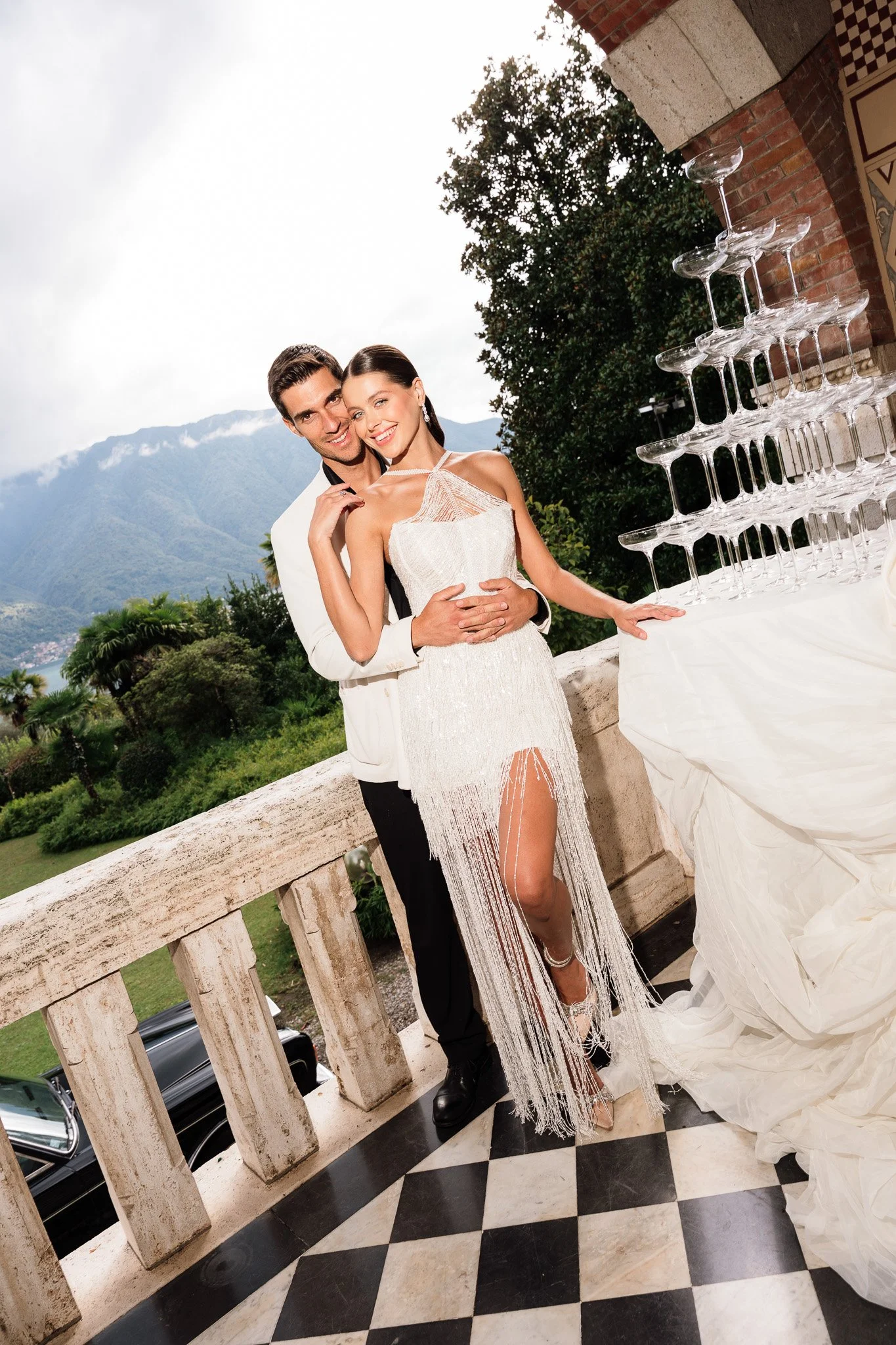 Ein glückliches Paar beim Hochzeitsfest, stehendes nebeneinander mit Bergen im Hintergrund, neben einem Turm aus Weingläsern, auf einem Balkon mit schwar-weißem Fliesenboden.