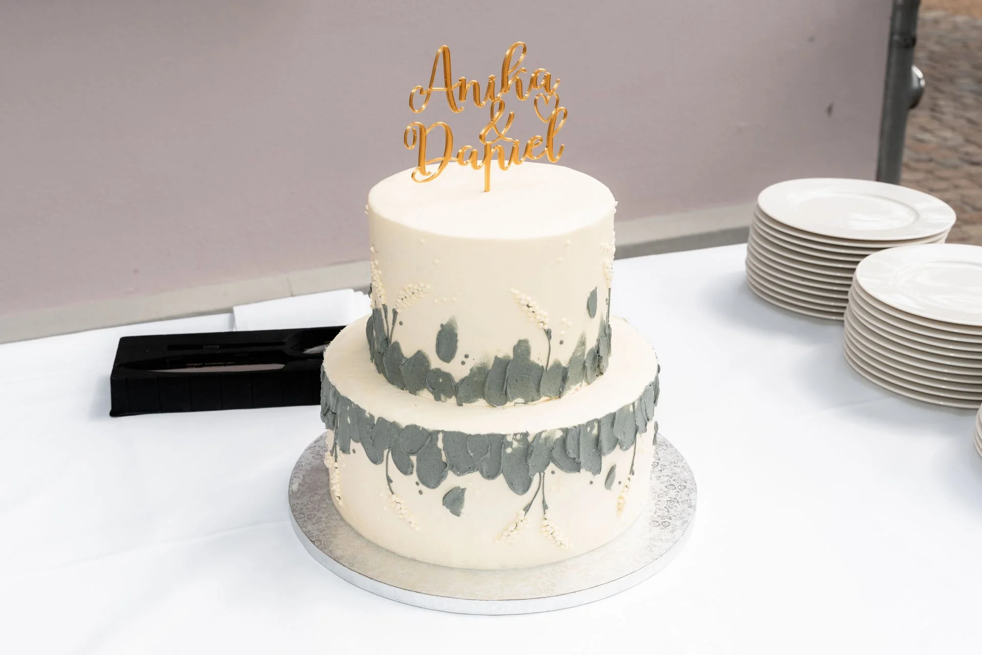 Zweistöckige Hochzeitstorte mit weißen und grauen Dekorationen, auf einem silbernen Tisch, mit einer goldenen Tortenfigur mit den Namen Anka und Daniel.