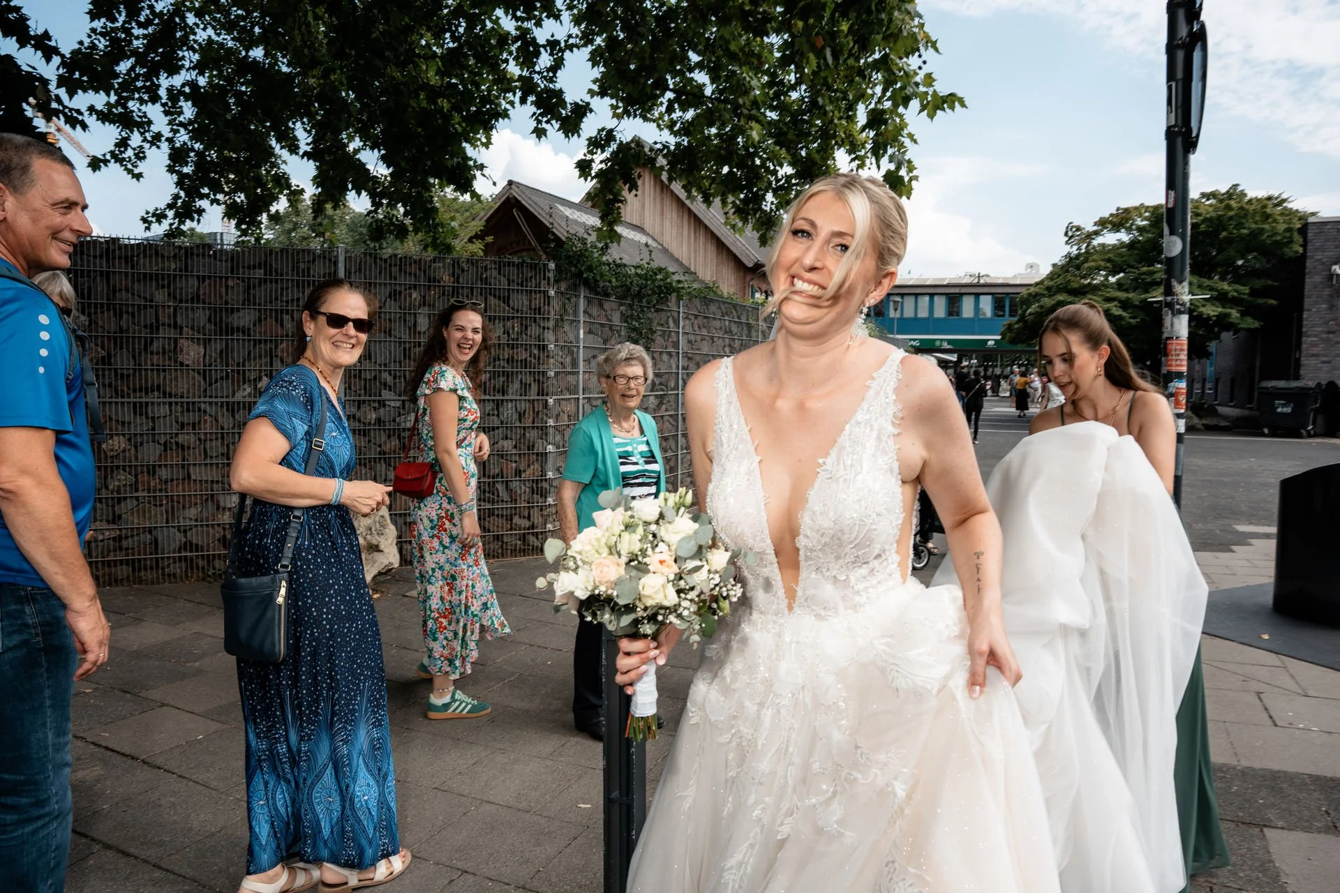 Eine Braut mit einem emotionalen Gesichtsausdruck, umgeben von lachenden und fröhlichen Menschen bei einer Hochzeit im Freien.