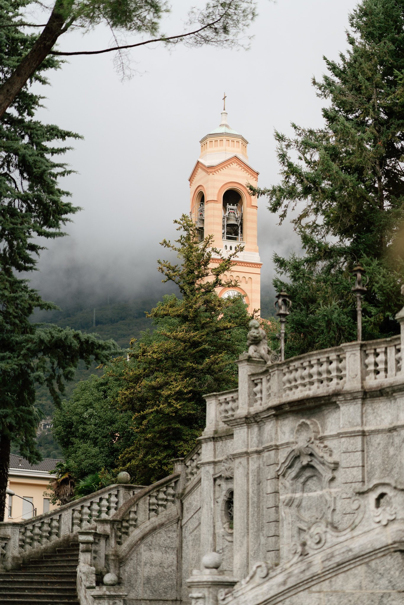 Ein rosafarbener Kirchturm mit Glocken, umgeben von Bäumen und einer Steinmauer im Vordergrund, mit Wolken und Bergen im Hintergrund.