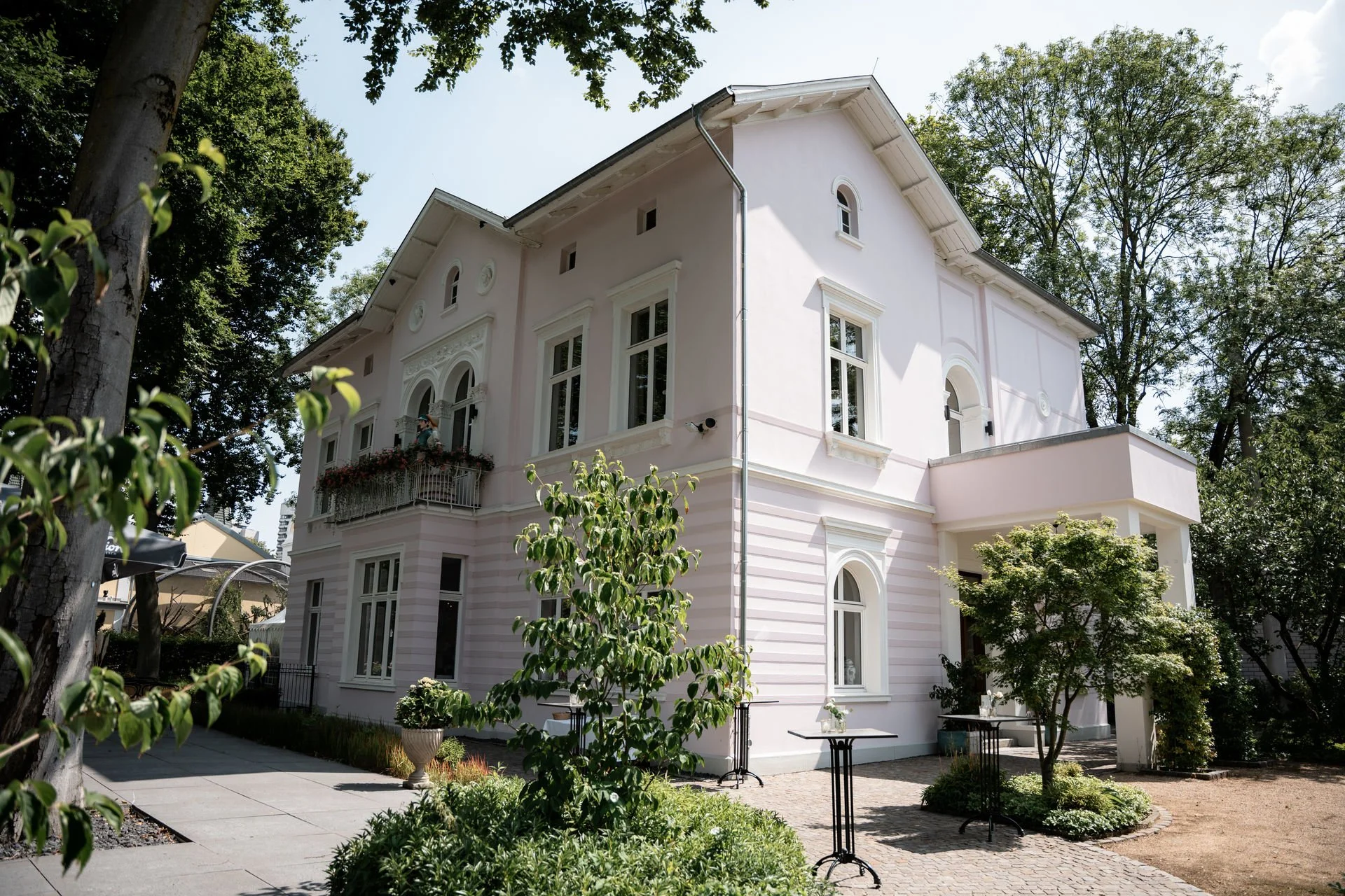 Ein großes, rosa farbenes Haus mit weißen Fenstern und Balkon, umgeben von grünen Bäumen und Pflanzen, bei sonnigem Wetter.