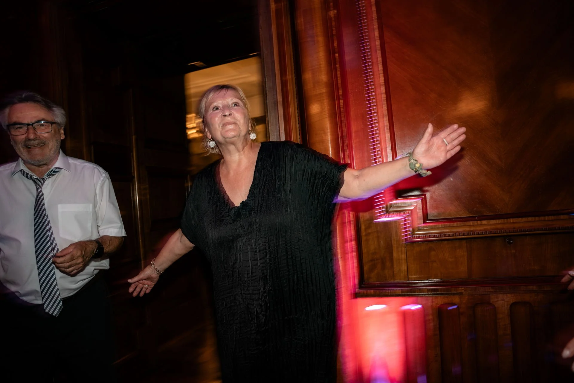 Eine ältere Frau trägt ein schwarzes Kleid und gestikuliert mit den Armen während einer Feier oder Veranstaltung, neben einem Mann in weißem Hemd und Krawatte im dunklen Raum mit Holzvertäfelung.