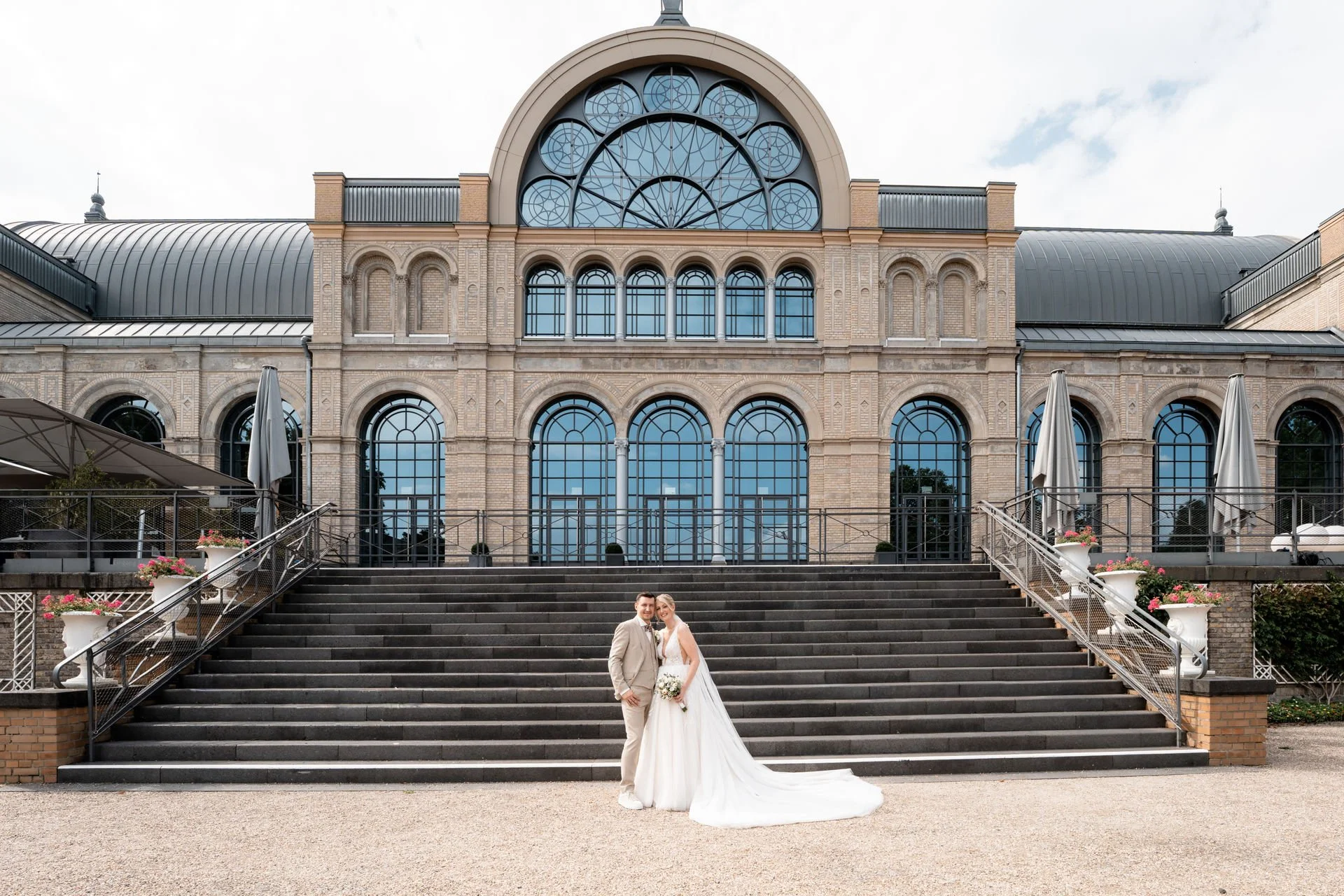Ein Brautpaar vor einem historischen Gebäude mit großen, bogenförmigen Fenstern und Treppe, die hoch zum Eingang führt.