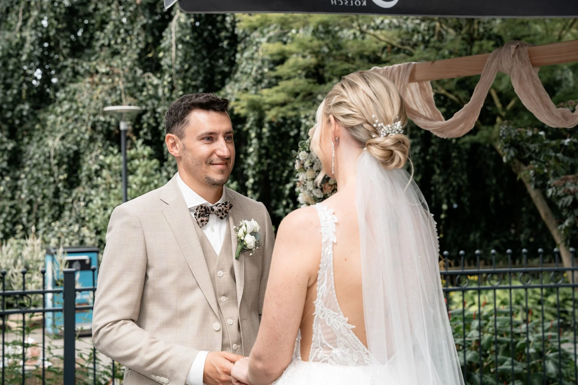 Ein Mann und eine Frau bei einer Hochzeit im Freien, die sich gegenseitig die Hände halten, vor einem grünen Hintergrund mit einem dekorativen Bogen aus Stoff.