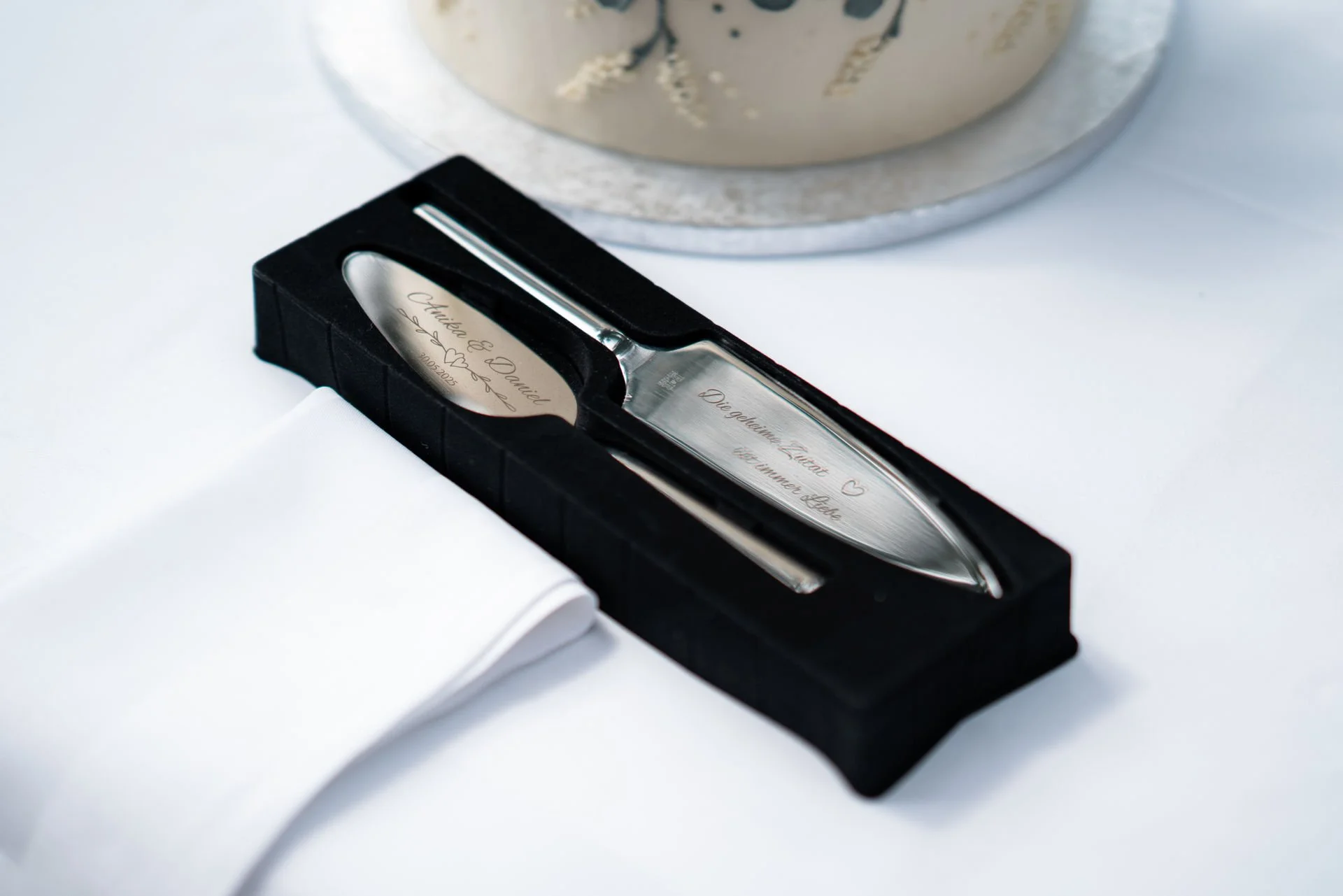 Ein er Silberlöffel in einer schwarzen Verpackung mit eingravierter Botschaft, daneben eine weiße Serviette, auf einem weißen Tisch, mit einem silbernen Kuchen im Hintergrund. Die Gravur auf dem Löffel lautet 'Christina & Daniel, 25.09.2023.'