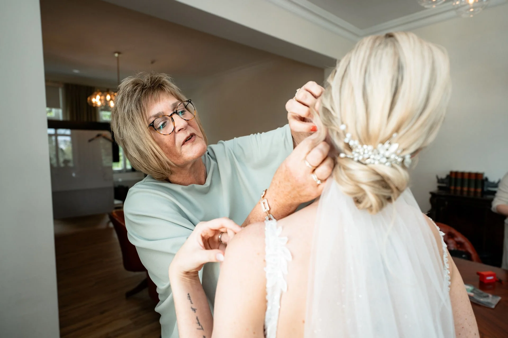 Eine ältere Frau richtet die Hochsteckfrisur einer jungen Braut. Die Braut trägt einen Schleier und eine Perlenbrosche im Haar, während die Frau konzentriert arbeitet.