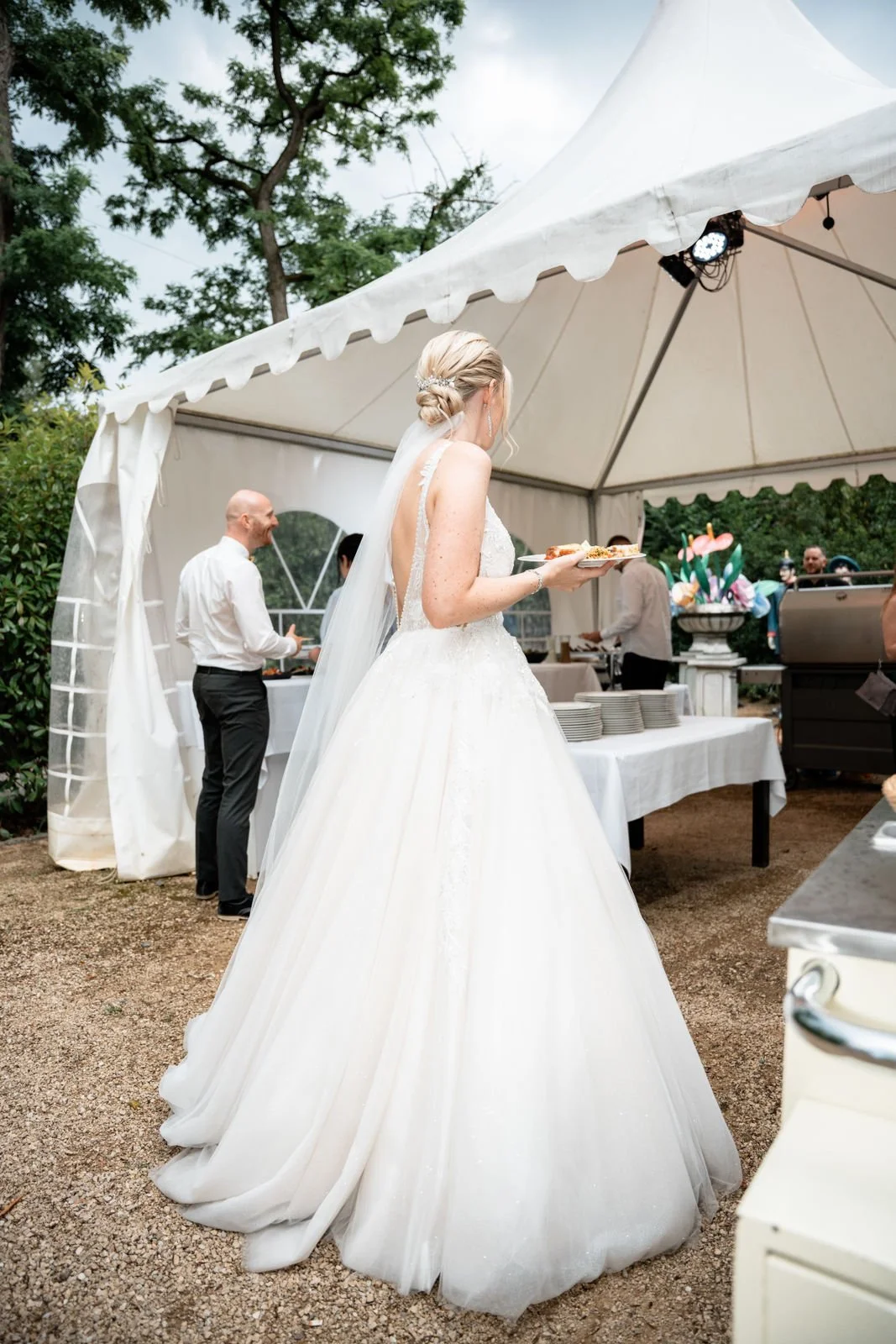 Braut in weißem Hochzeitskleid mit langem Saum und Schleier, die einen Teller mit Essen hält, bei einer Gartenhochzeit unter einem weißen Zelt, im Hintergrund weitere Personen beim Essen, und attraktive Dekorationen mit bunten Blumen.