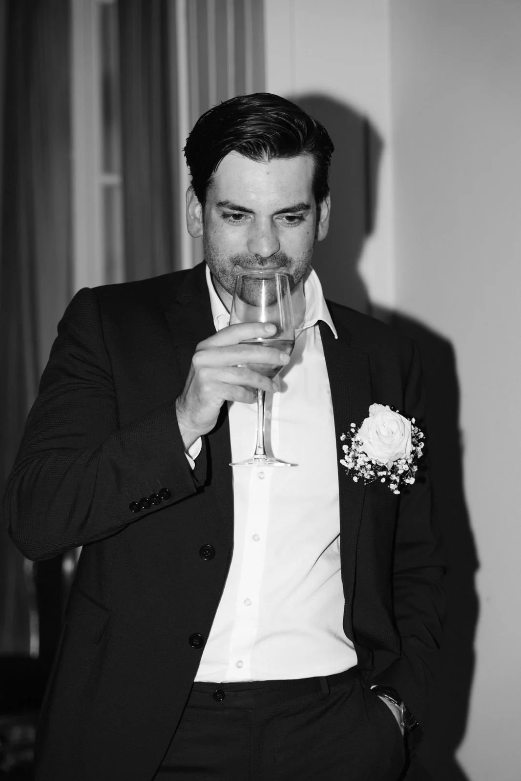 Schwarz-weiß-Foto eines Mannes in formeller Kleidung, der ein Weinglas hält und bei einer Feier, möglicherweise einer Hochzeit, steht.