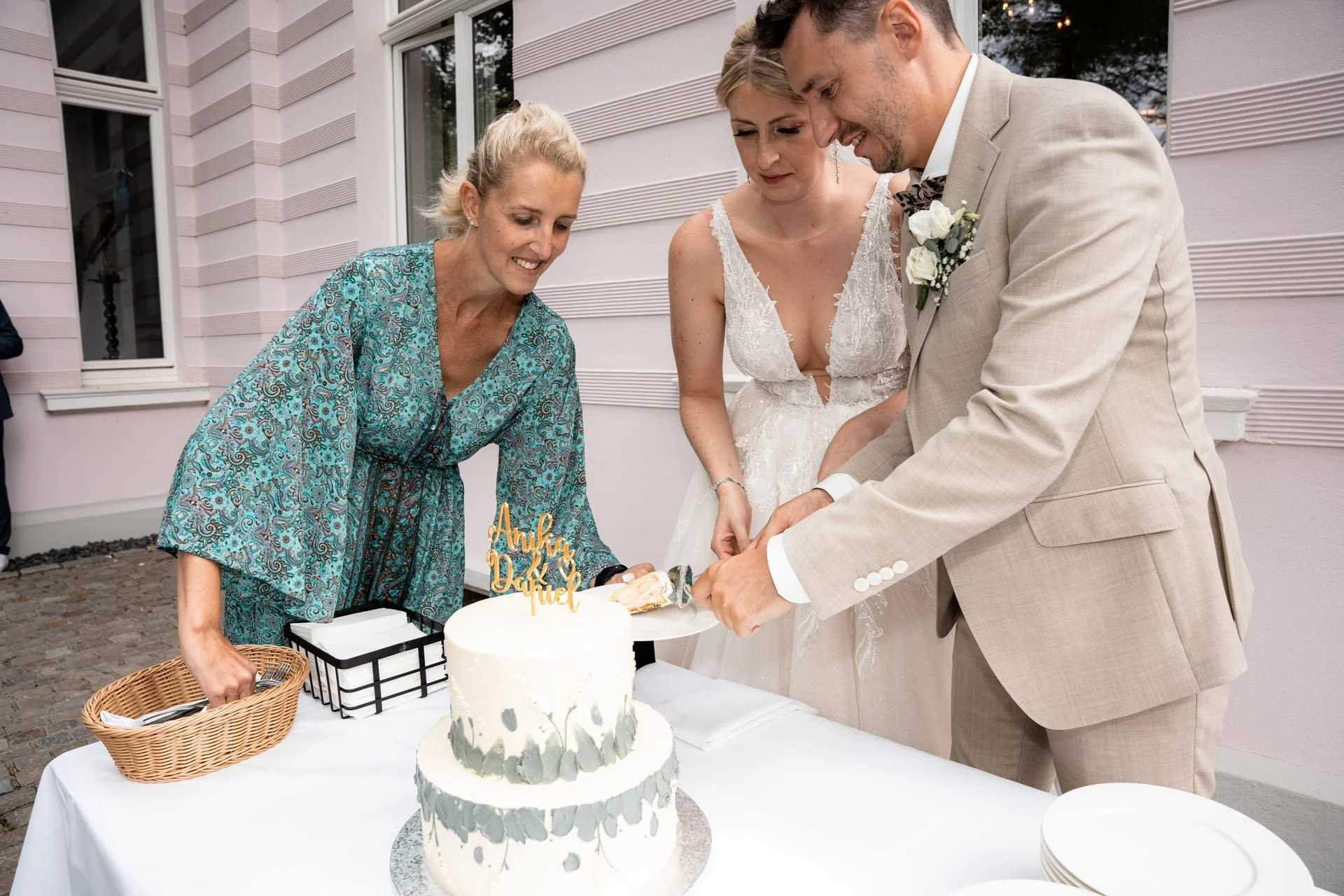 Hochzeitspaar schneidet die Hochzeitstorte traditionell mit Hilfe einer Frau. Das Brautpaar steht vor einem weißen Tisch mit Torte, im Hintergrund ein pinkes Haus.