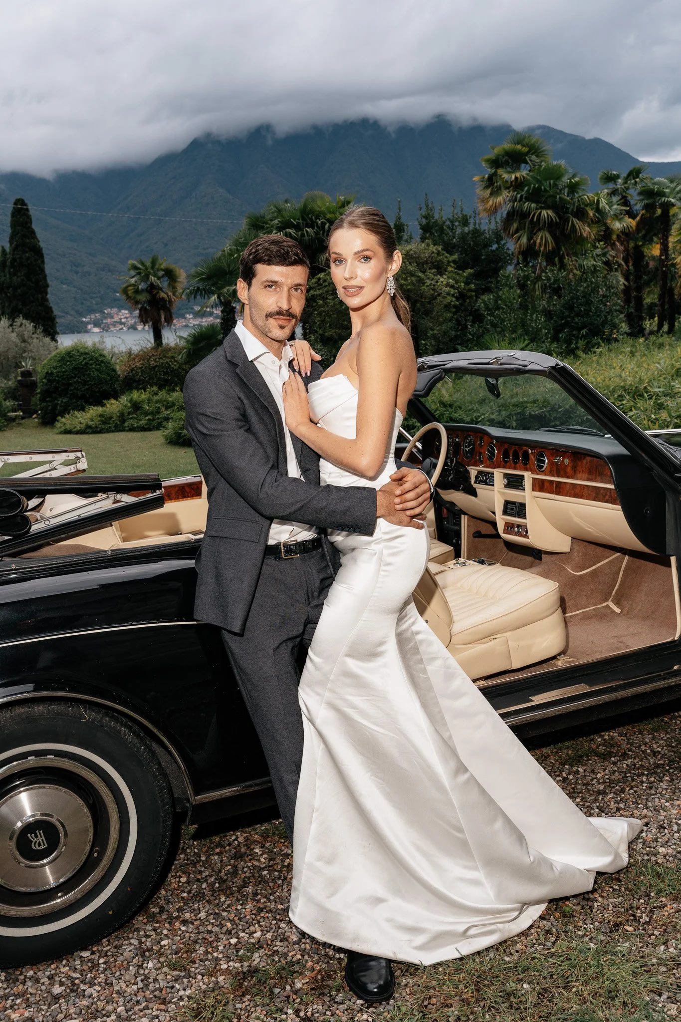 Ein Brautpaar in Hochzeitskleidung posiert vor einem offenen Oldtimer-Cabriolet, im Hintergrund sind Berge und bewölkter Himmel zu sehen.