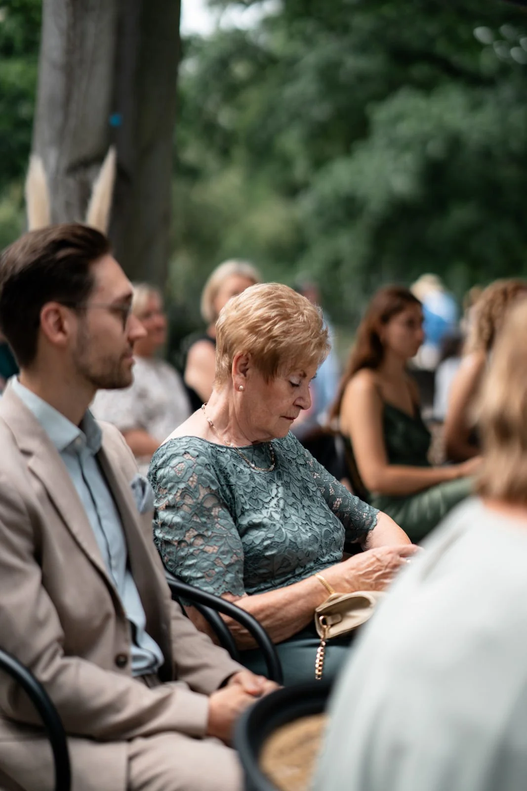 Menschen sitzen im Freien bei einer Veranstaltung, viele konzentrieren sich auf etwas außerhalb des Bildes, einige sind in formeller Kleidung, umgeben von Bäumen und Grün.