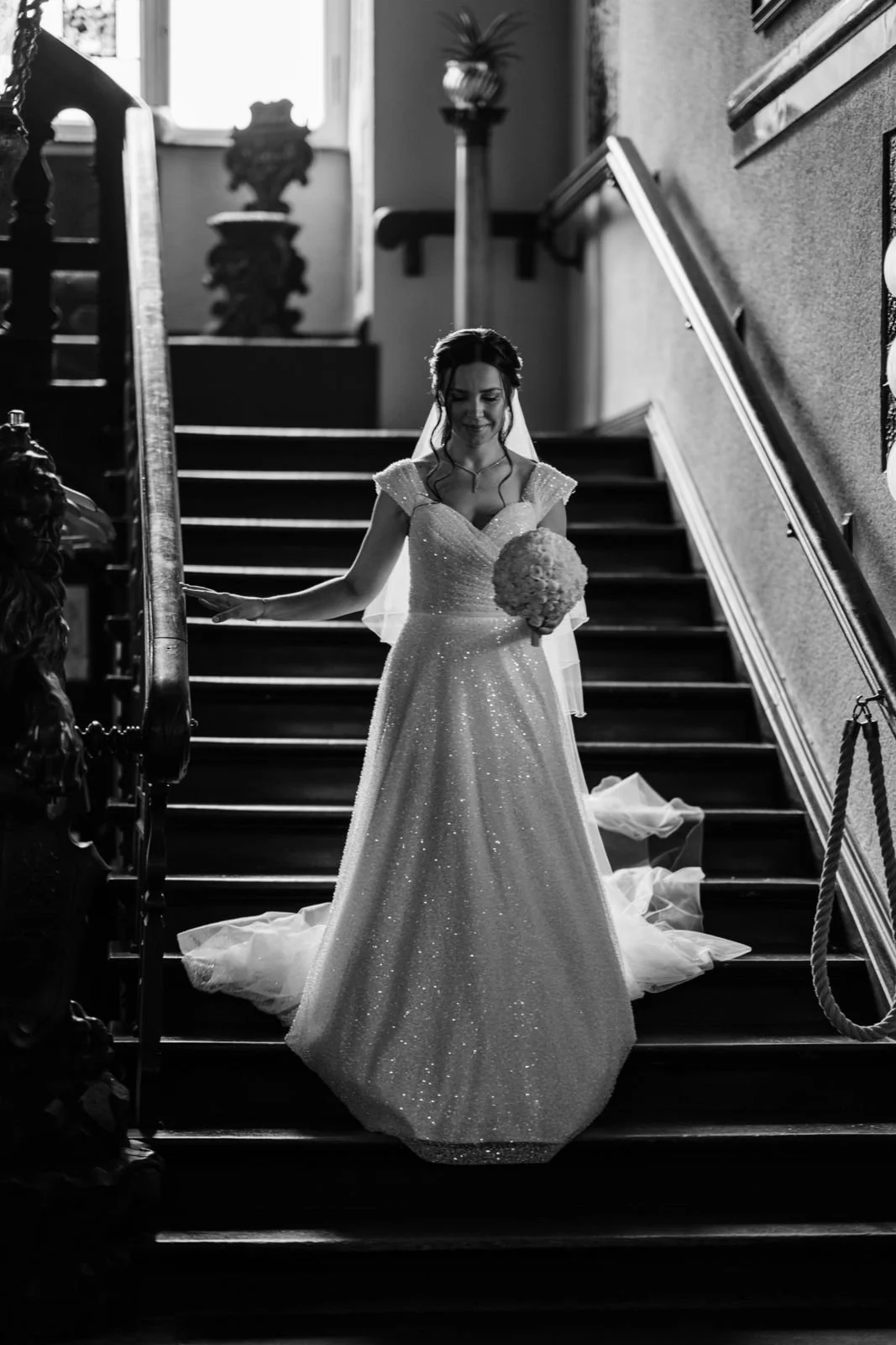 Braut in einem schimmernden Hochzeitskleid mit Schleier, die auf einer Treppe steht und einen Brautstrauß hält, während sie nach unten blickt.