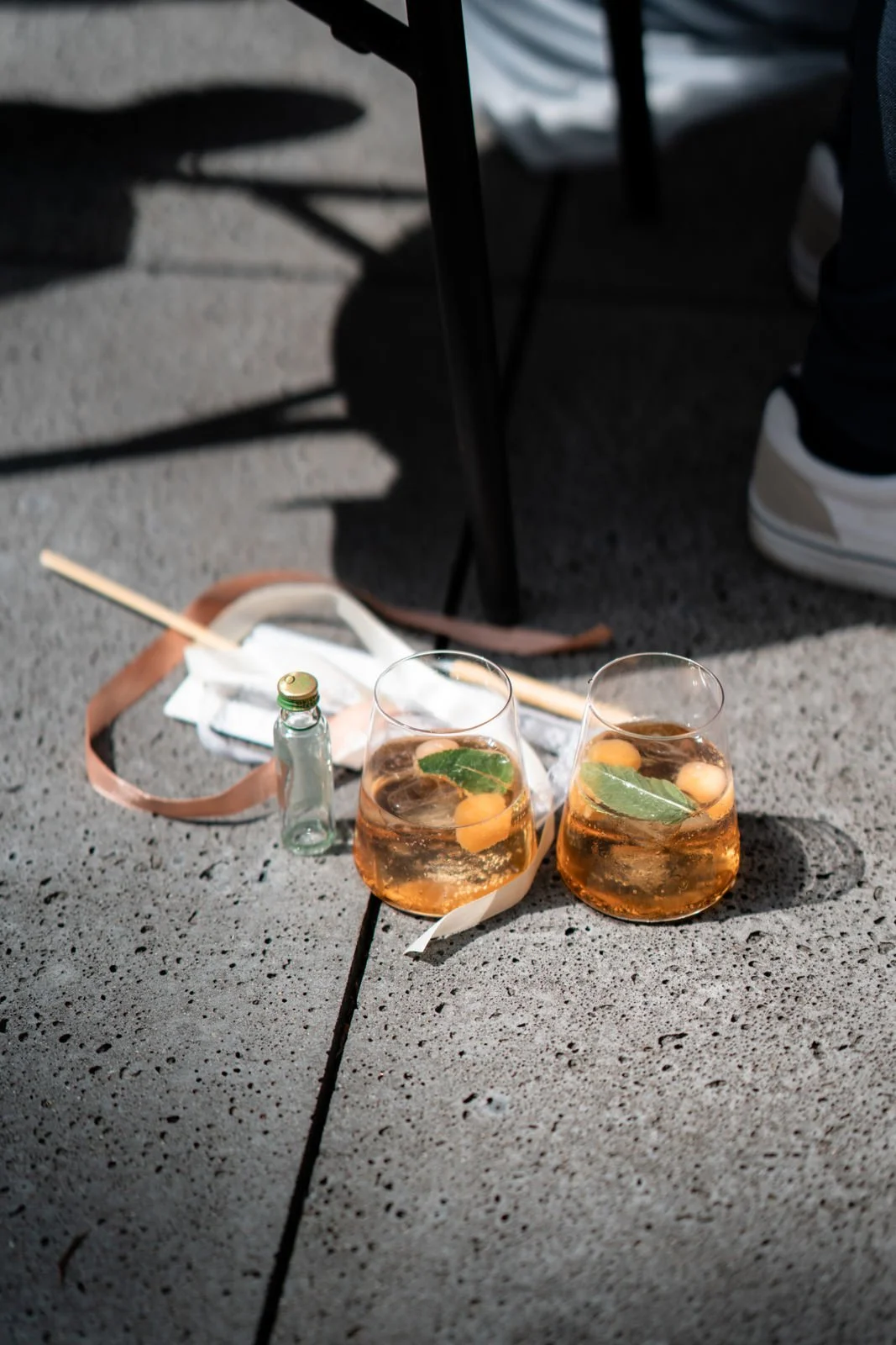 Zwei Gläser mit Whisky, Limetten und Ohrstäbchen auf einem Gehweg, daneben eine kleine Glasflasche und eine neutrale Maske.