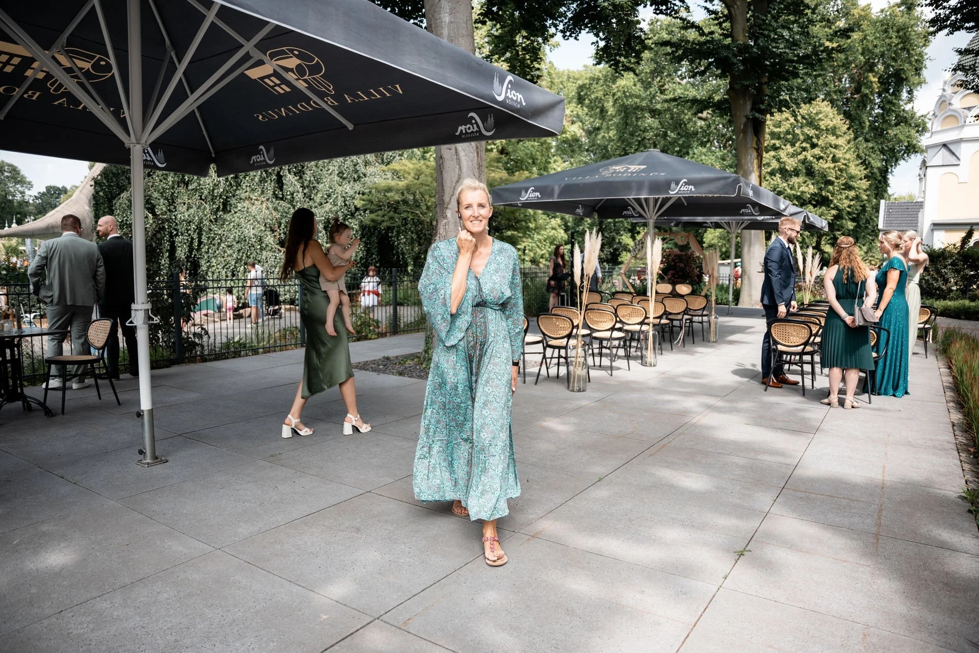 Eine Gruppe von Menschen bei einer Feier im Freien, einige stehen, andere sitzen, mit großen Sonnenschirmen und Bäumen im Hintergrund.
