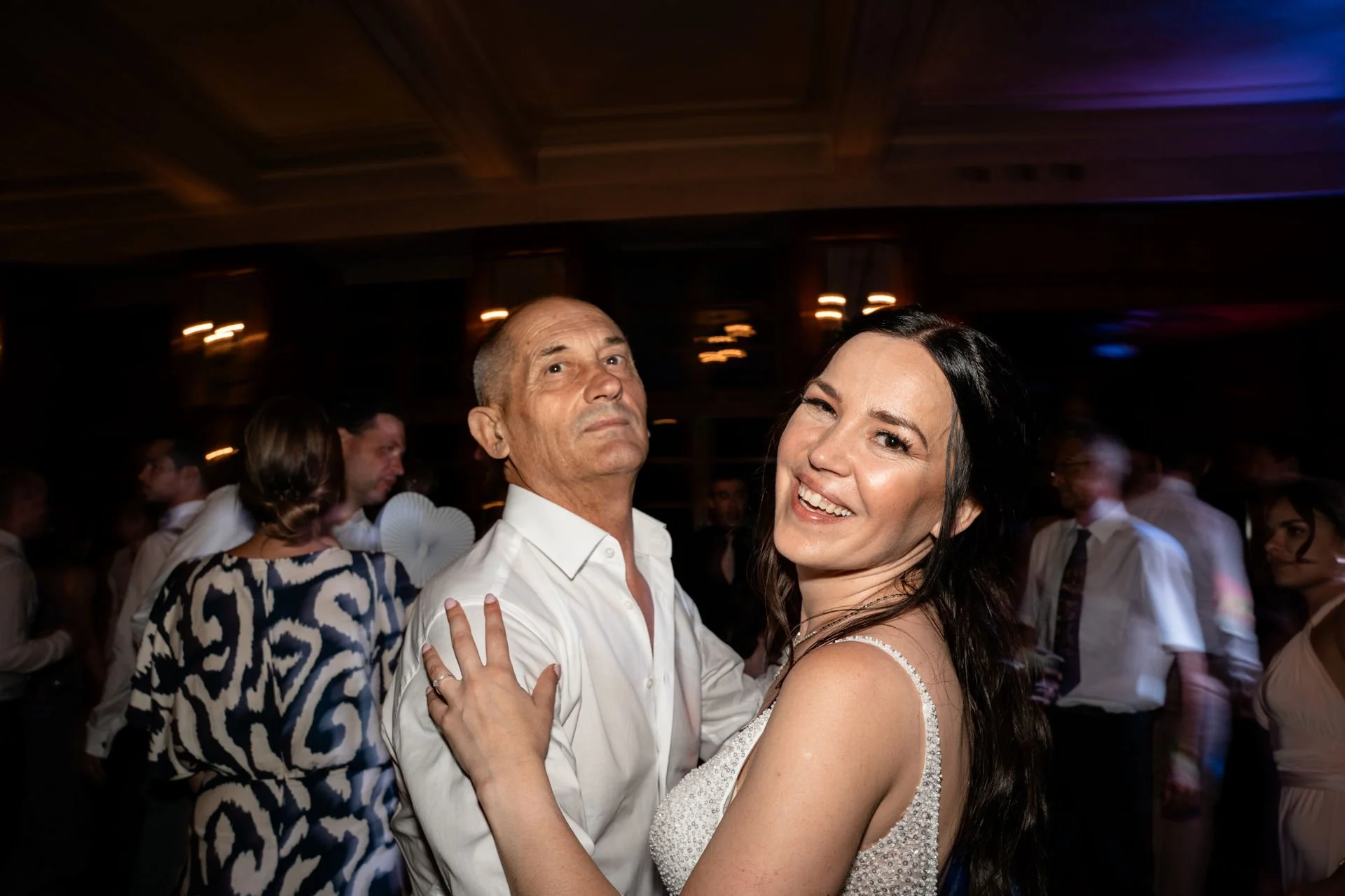 Zwei Menschen tanzen auf einer Party. Der ältere Mann trägt ein weißes Hemd, die junge Frau trägt ein glitzerndes Kleid. Im Hintergrund weitere Gäste.