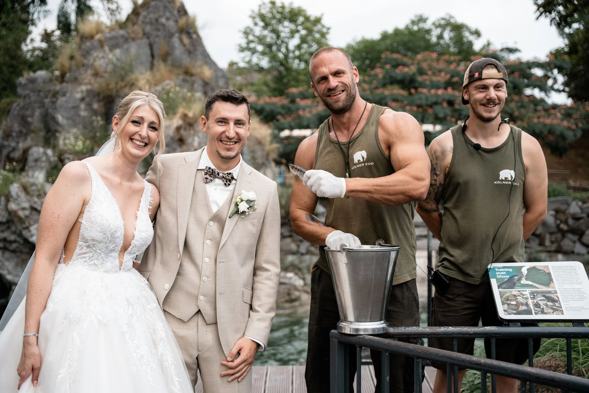 Eine Hochzeitsgesellschaft steht neben einem Tierpfleger mit einer Fisch in der Hand in einem Zoo-Garten.