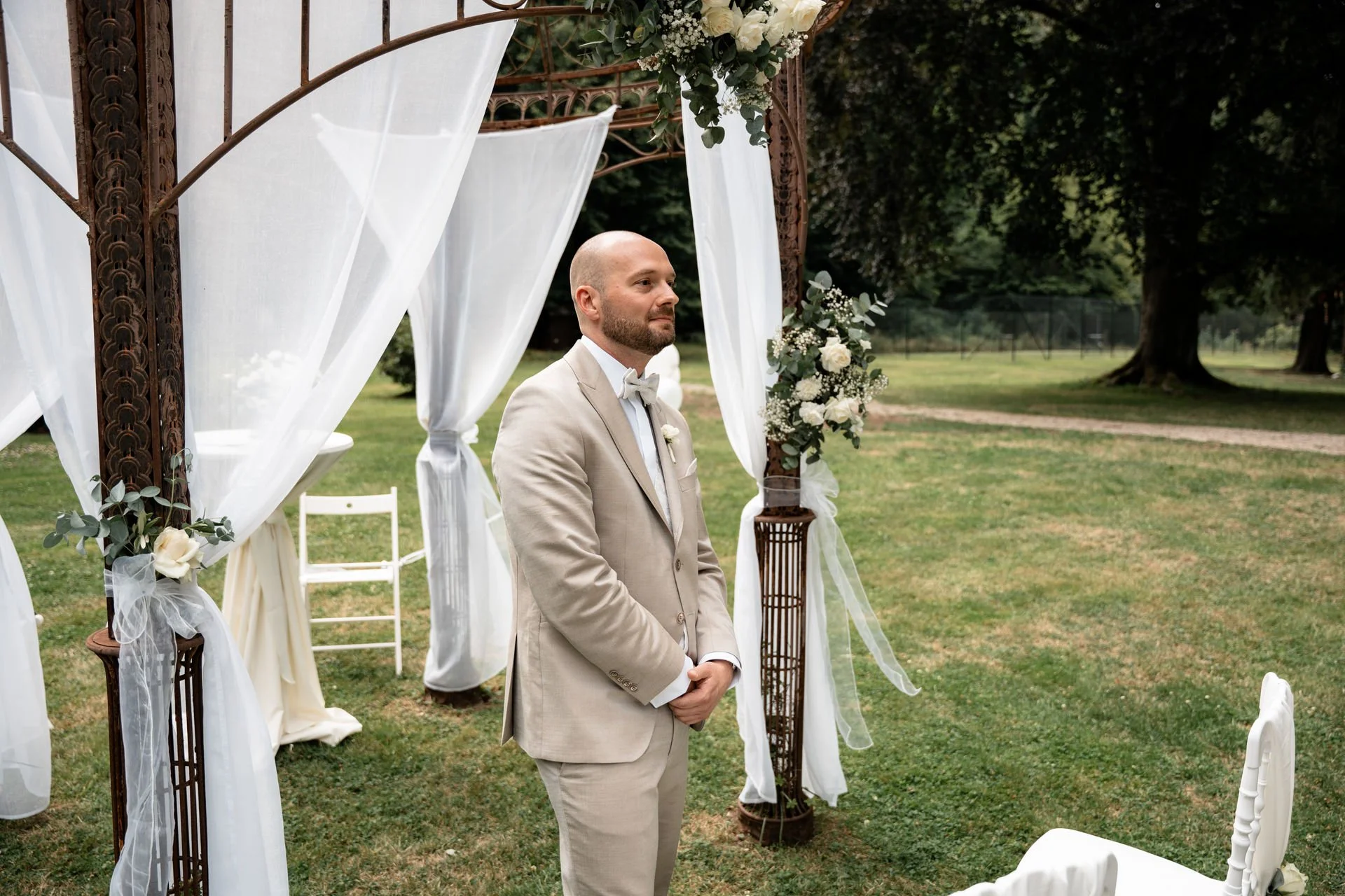 Ein Mann in einem hellen Anzug steht an einem Hochzeitsschaukel im Freien, umgeben von weißen Tüchern und Blumen, während er auf die Zeremonie wartet.