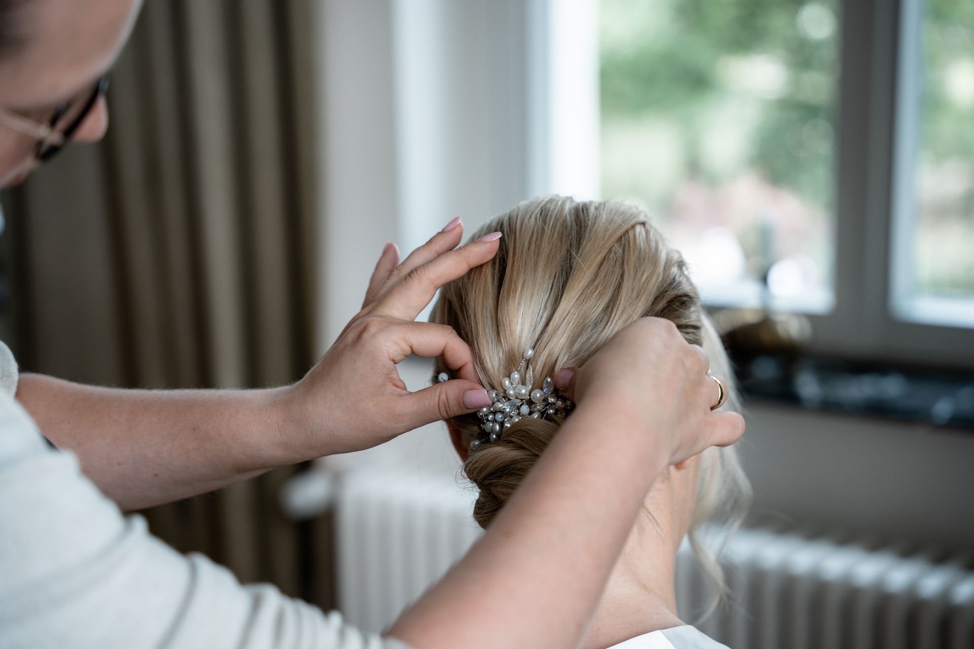 Eine Frau wird von einer Stylistin frisiert, die an ihrem Haar arbeitet, das mit einem eleganten, perlenden Haarstyling-Haaraccessoire geschmückt ist, in einem hell erleuchteten Raum mit Fenster im Hintergrund.