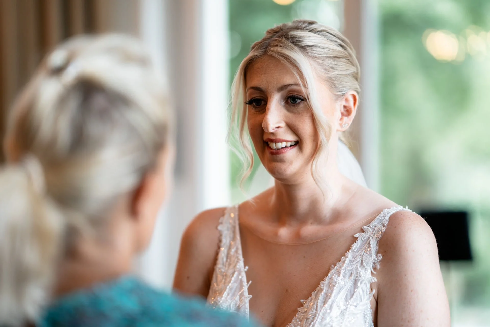 Zwei Frauen, eine Braut und eine ältere Frau, im Gespräch in einem hellen Raum mit großen Fenstern im Hintergrund.