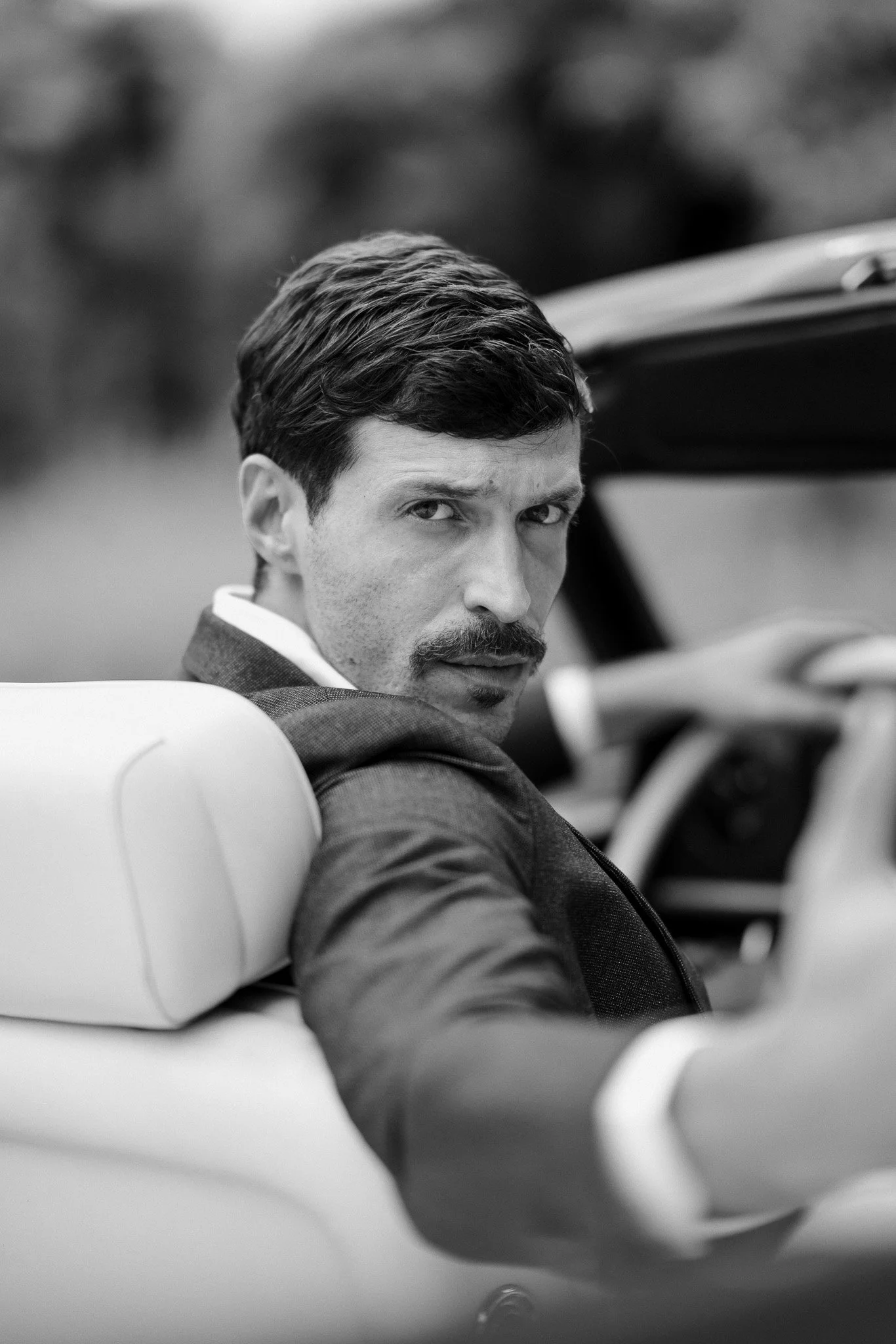 Schwarz-weißer Foto eines Mannes mit dunklem Haar, Schnurrbart und Anzug in einem Cabrio, der direkt in die Kamera schaut.