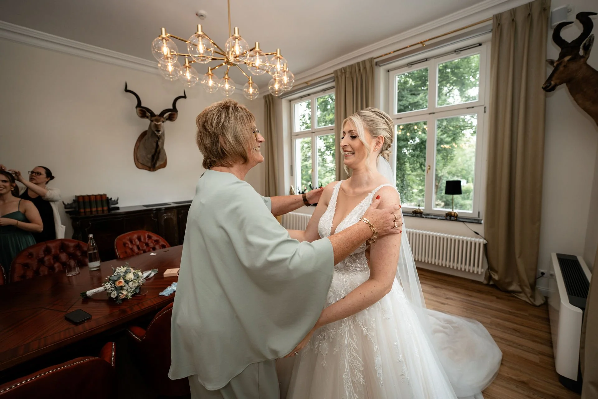 Hochzeit: Braut und Mutter umarmen sich in einem hellen Raum mit großen Fenstern, Dekorationen und einem Tisch mit Blumen.