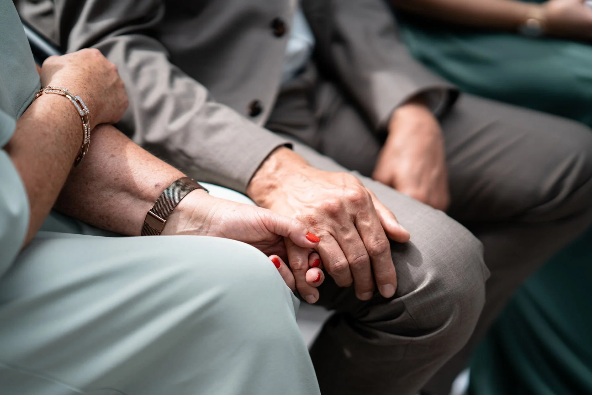 Nahaufnahme von zwei älteren Händen, die sich halten, auf einem Sitz im Zentrum eines Treffens. Die Hände sind voneinander umschlossen und zeigen ein Gefühl von Unterstützung und Verbundenheit.