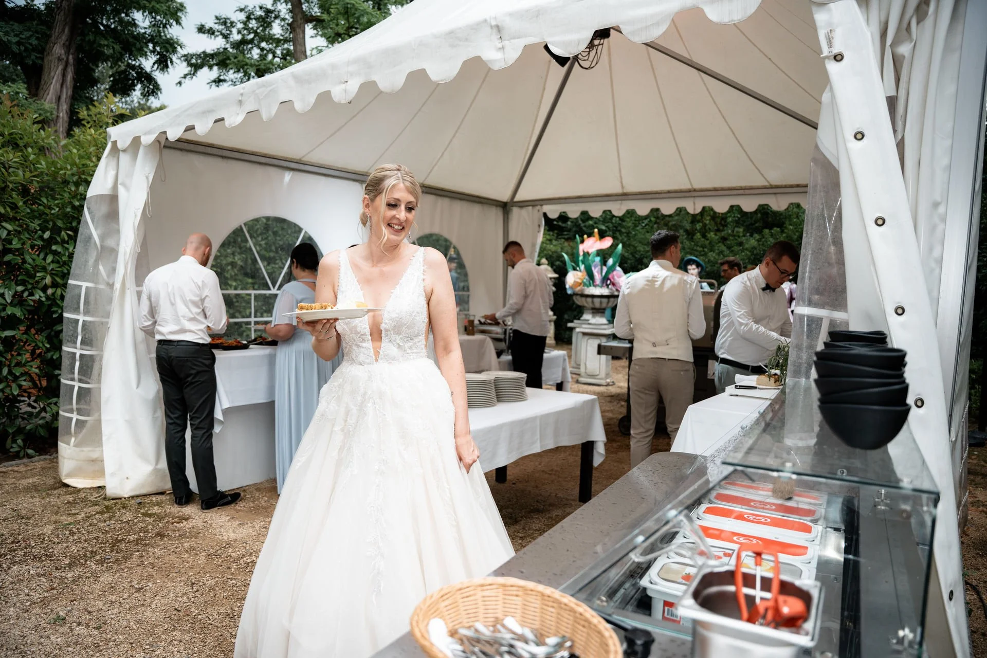 Braut in Hochzeitskleid beim Buffet, umgeben von Hochzeitsgästen in formeller Kleidung, im freien, unter einem weißen Zelt, mit Eiscreme-Station im Vordergrund.