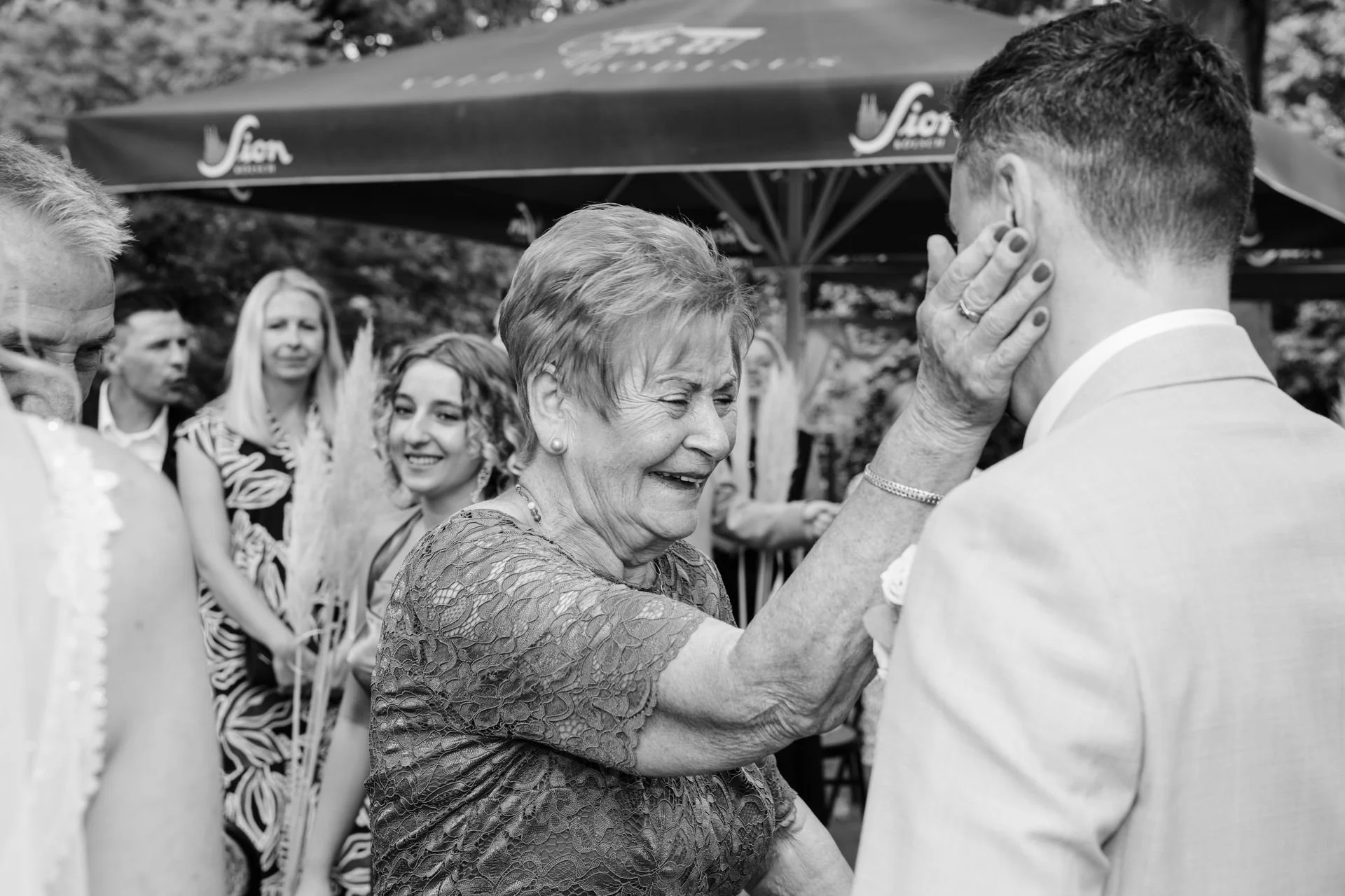 Eine ältere Frau lächelt, während sie einem jungen Mann im Anzug mit der Hand ins Gesicht fasst, bei einer Feier im Freien unter einem Sonnenschirm.
